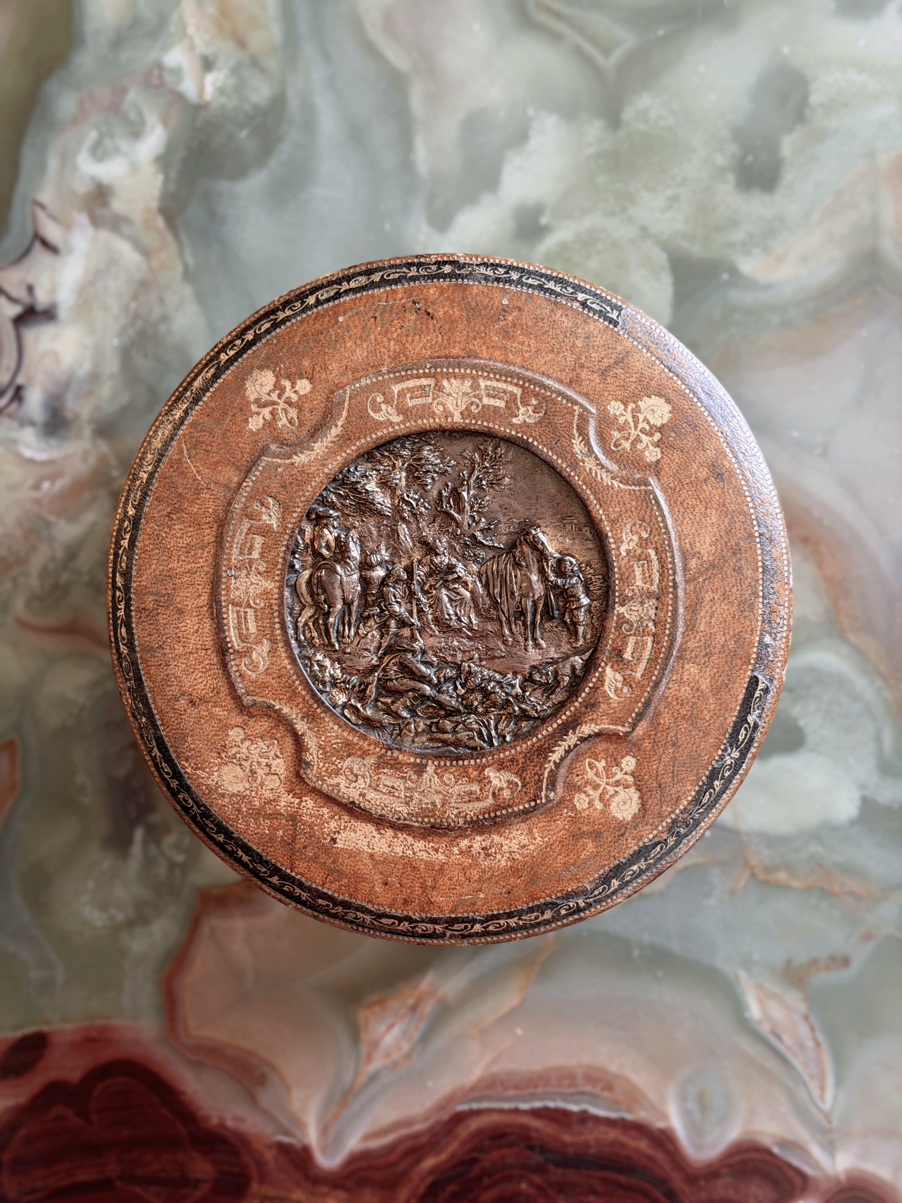Antique French Grand Tour leather vanity box from the early 20th century, showing a green tooled Morocco leather exterior with a central cast bronze repoussé plaque depicting a classical pastoral garden party scene, opened to reveal a blush-pink ruched silk faille interior and original steel barrel key.