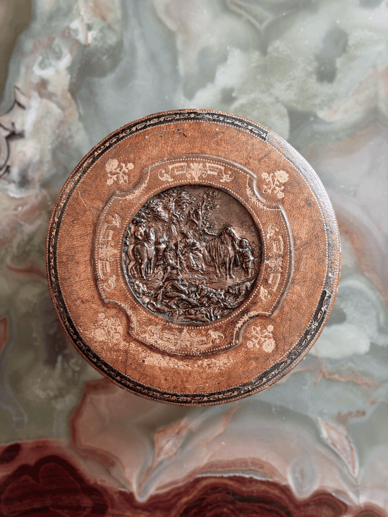 Antique French Grand Tour leather vanity box from the early 20th century, showing a green tooled Morocco leather exterior with a central cast bronze repoussé plaque depicting a classical pastoral garden party scene, opened to reveal a blush-pink ruched silk faille interior and original steel barrel key.