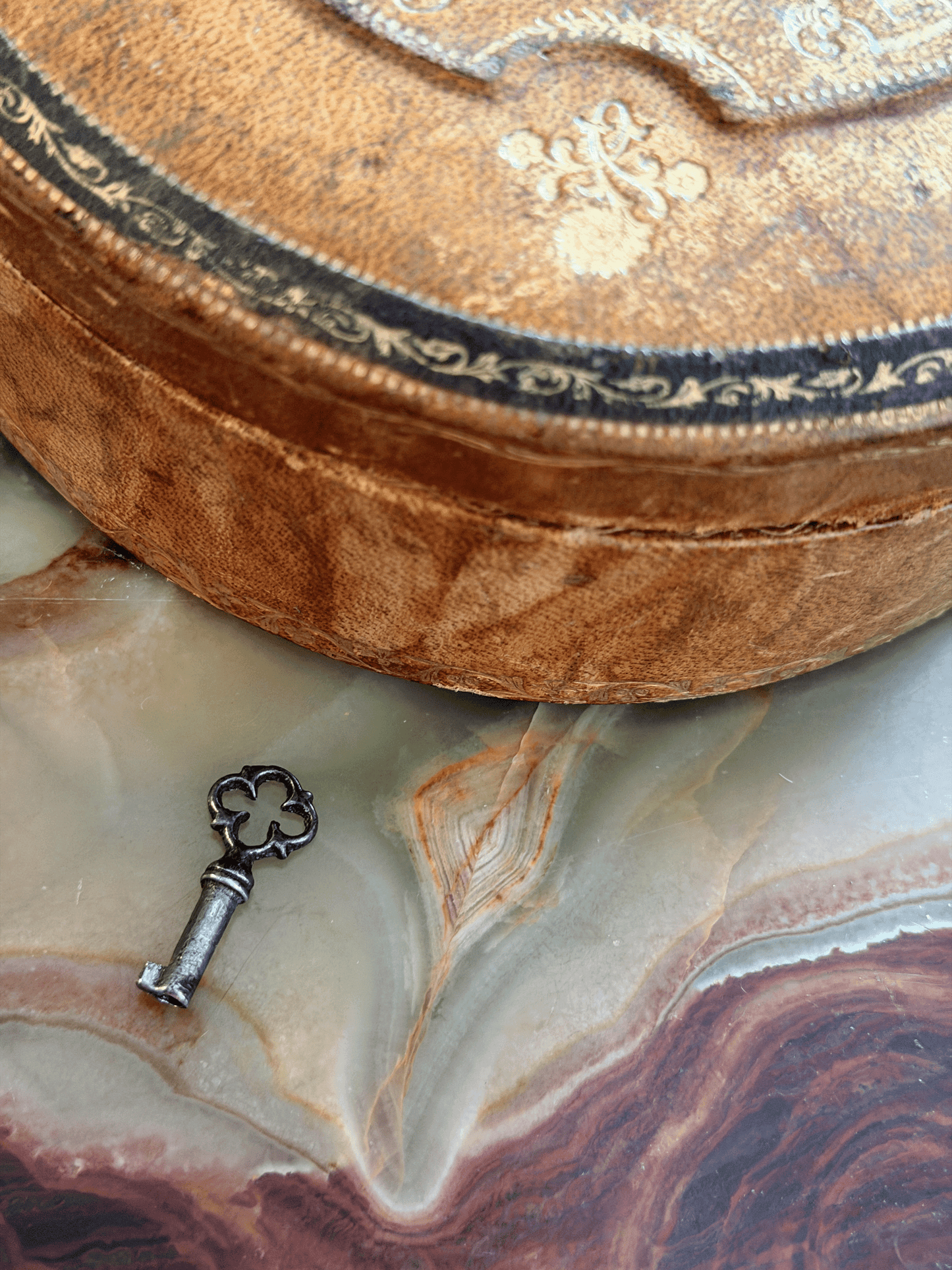 A close-up of an antique oval wooden box with ornate gold-toned floral engravings and a black decorative border, resting on a polished agate slab next to a small vintage key.