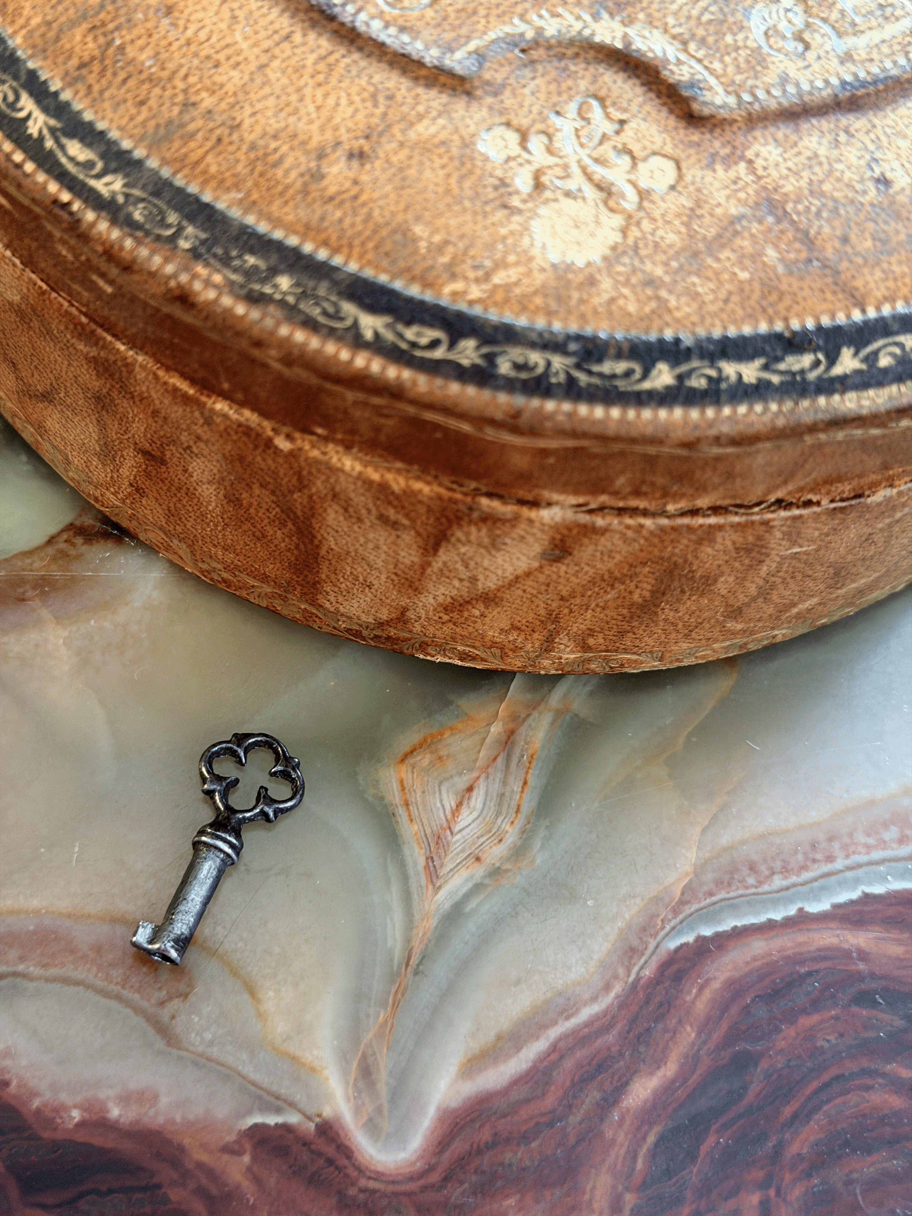 A close-up of an antique oval wooden box with ornate gold-toned floral engravings and a black decorative border, resting on a polished agate slab next to a small vintage key.