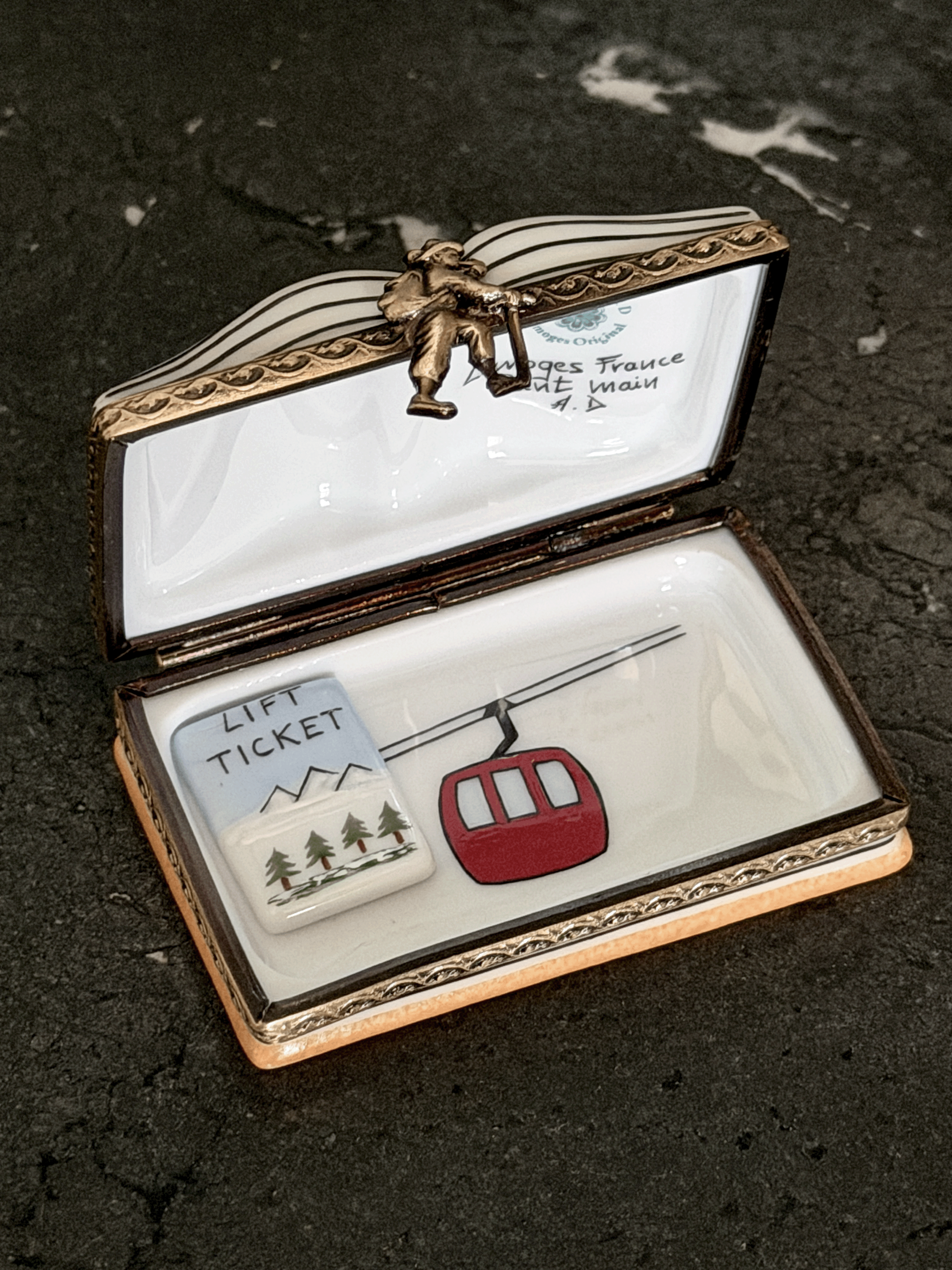 An open decorative trinket box with a white enamel interior and ornate gold-toned metal trim, featuring a small red cable car and a white lift ticket charm inside. The lid has a small figurine and handwritten text visible.