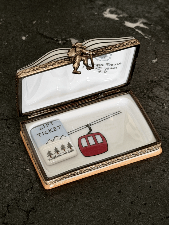 An open decorative trinket box with a white enamel interior and ornate gold-toned metal trim, featuring a small red cable car and a white lift ticket charm inside. The lid has a small figurine and handwritten text visible.