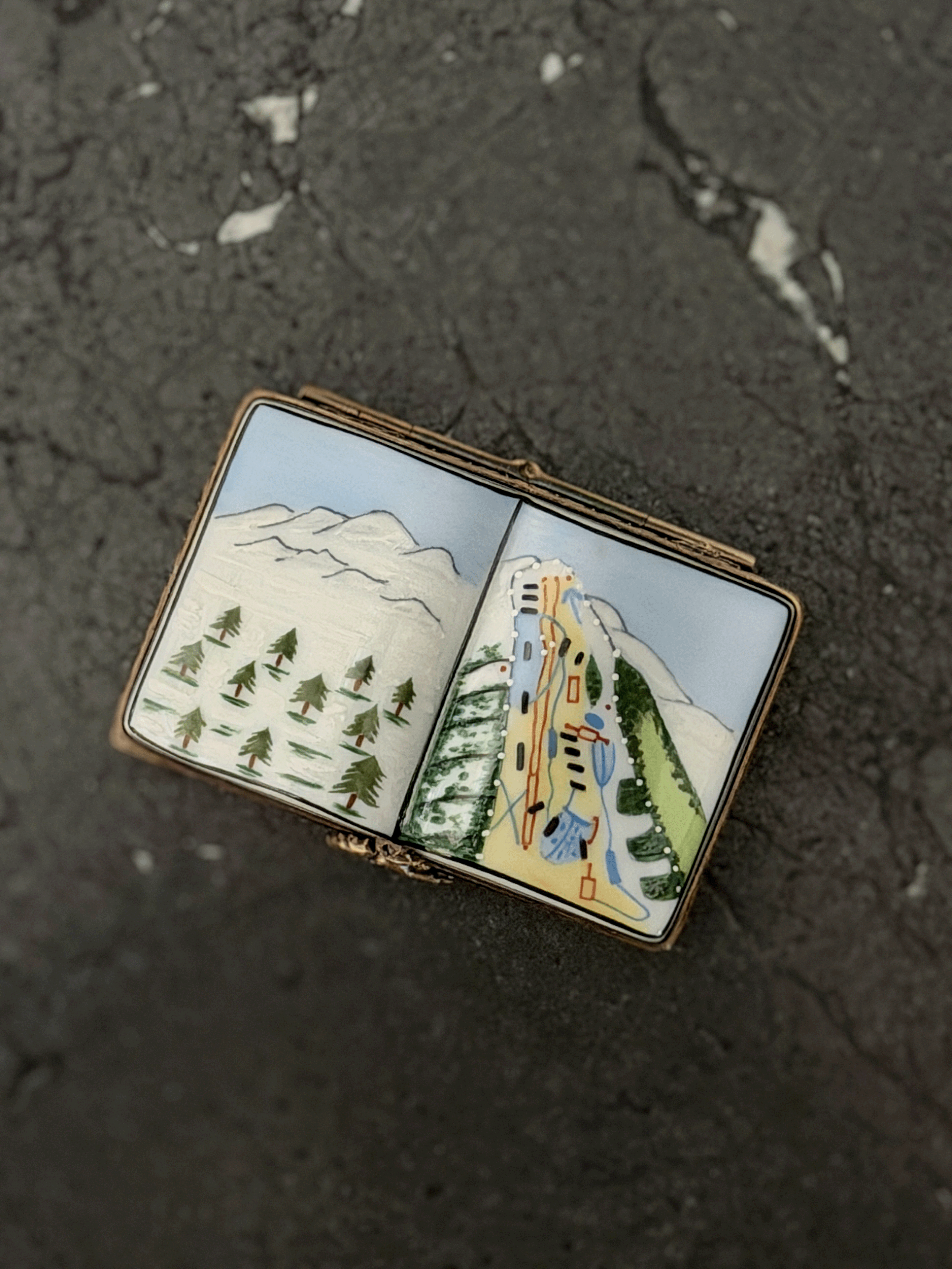 Hand-painted Rochard Limoges porcelain trinket box shaped like an open book, depicting a snow-capped alpine mountain peak on the left page and a colorful ski resort trail map with chairlift networks on the right page.