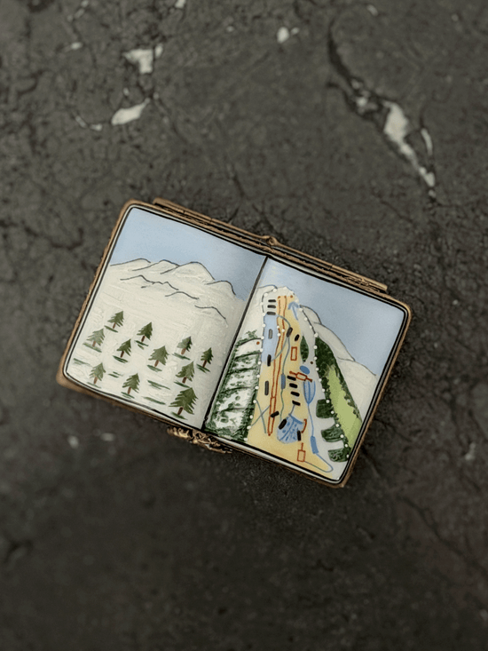 Hand-painted Rochard Limoges porcelain trinket box shaped like an open book, depicting a snow-capped alpine mountain peak on the left page and a colorful ski resort trail map with chairlift networks on the right page.