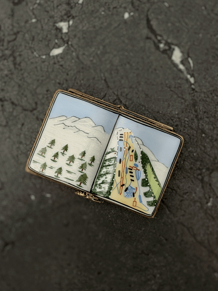 Hand-painted Rochard Limoges porcelain trinket box shaped like an open book, depicting a snow-capped alpine mountain peak on the left page and a colorful ski resort trail map with chairlift networks on the right page.