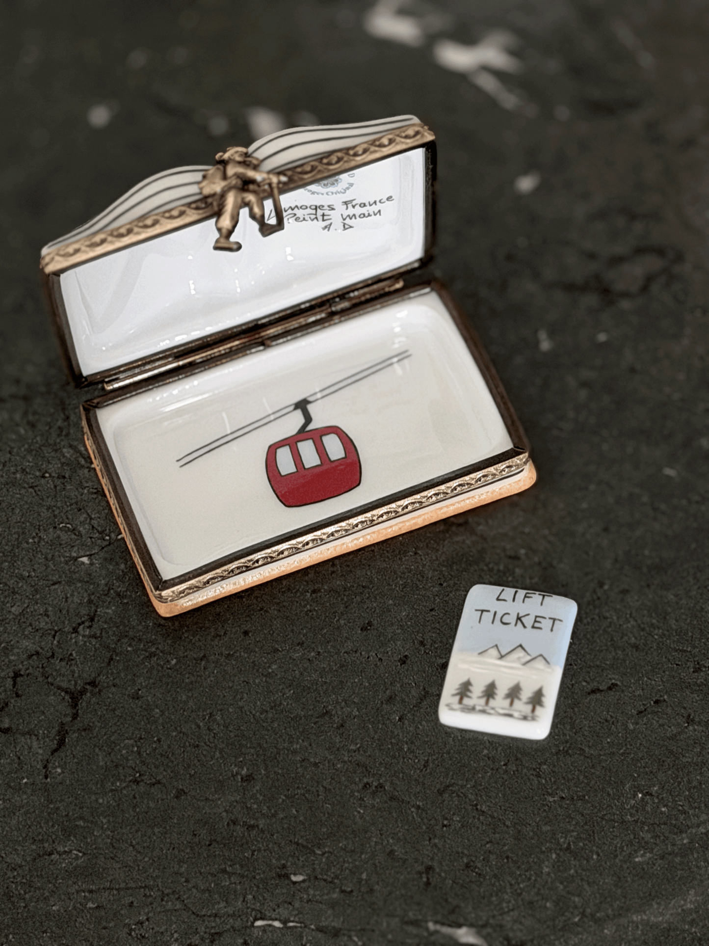An open Limoges-style enamel box with a lift ticket design, featuring a red cable car and a small matching 'Lift Ticket' card on a dark textured surface.