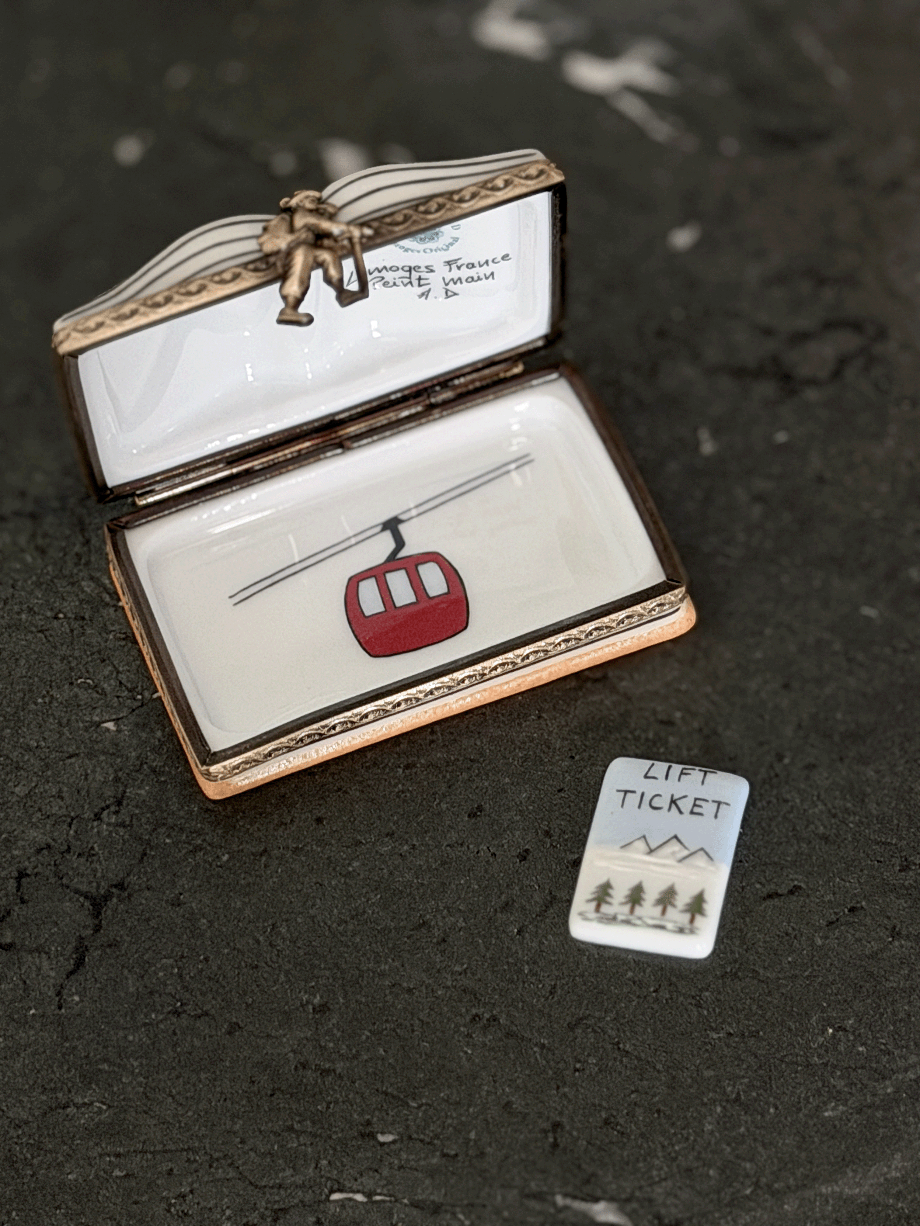 An open Limoges-style enamel box with a lift ticket design, featuring a red cable car and a small matching 'Lift Ticket' card on a dark textured surface.