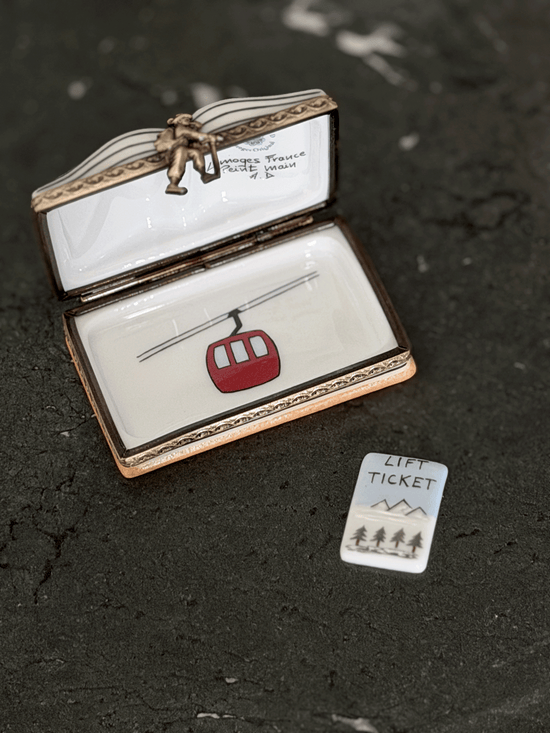 An open Limoges-style enamel box with a lift ticket design, featuring a red cable car and a small matching 'Lift Ticket' card on a dark textured surface.