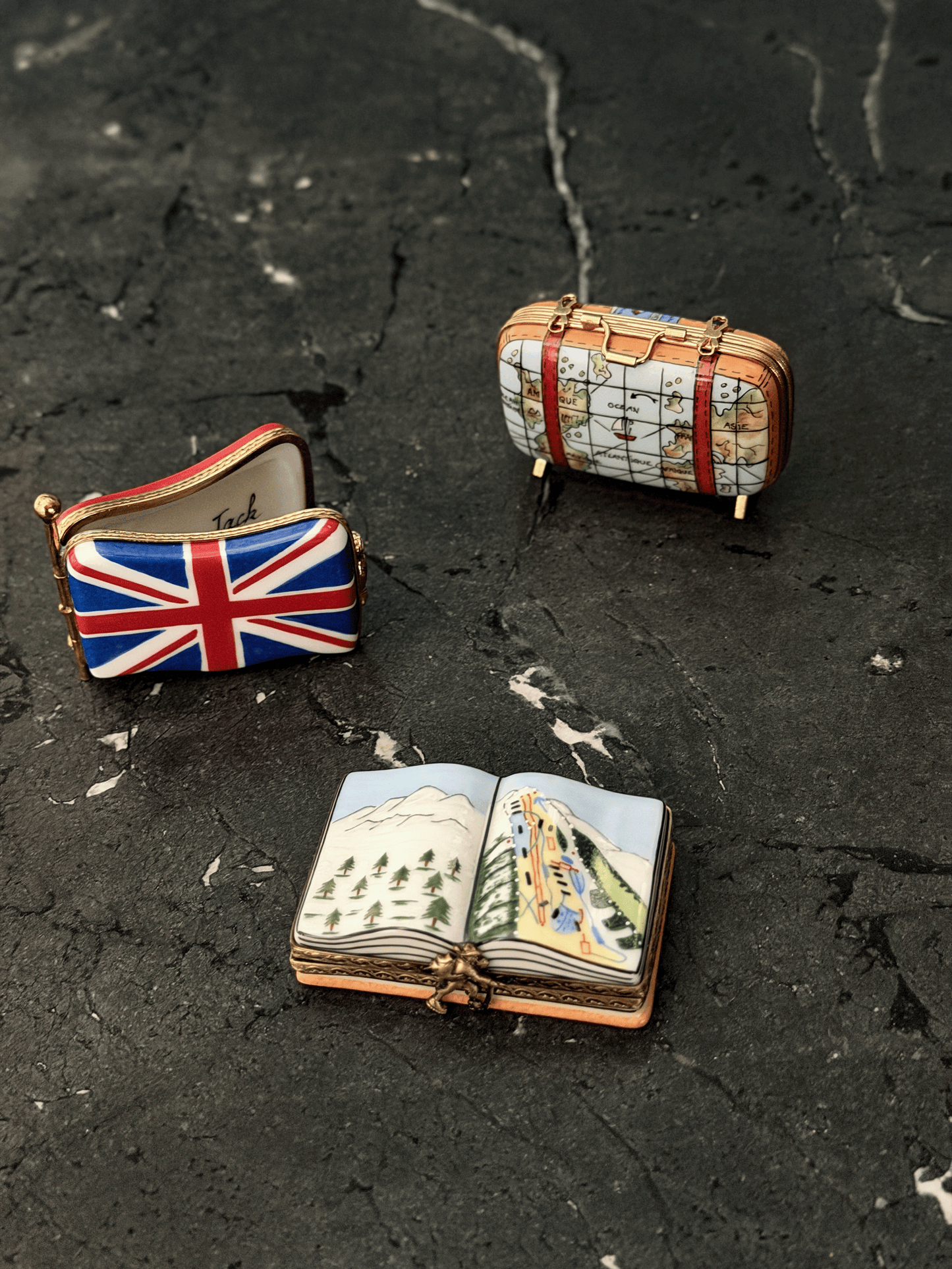 Three miniature travel-themed Limoges arranged on a dark textured surface: a small suitcase with a map print, a compact pouch featuring the Union Jack flag, and an open book charm with illustrated scenery.
