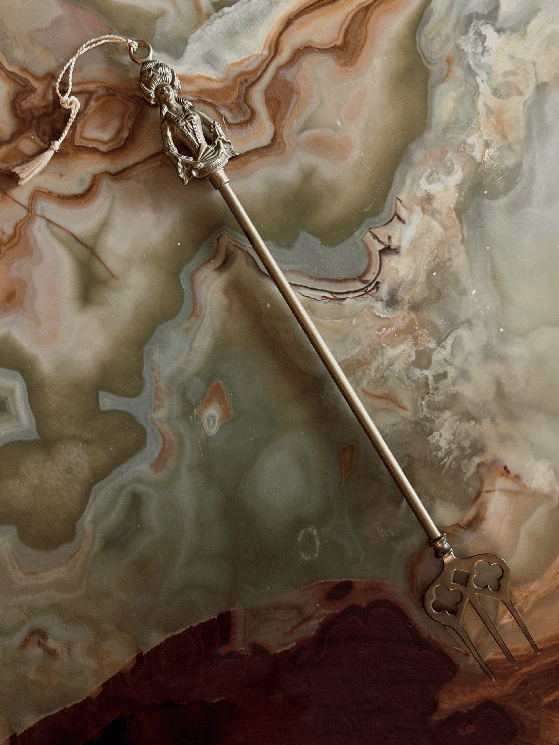 A vintage fireplace toasting fork, resting on a swirling cream and green onyx surface. The finial is sculpted into the shape of Madame de Pompadour in 18th-century French dress with a towering, high-swept hairstyle and a laced bodice. She holds a folded hand fan across her front that has the word POMPADOUR stamped clearly into it. A thin, braided cream string tassel is tied to a suspension ring at the very top of her head.