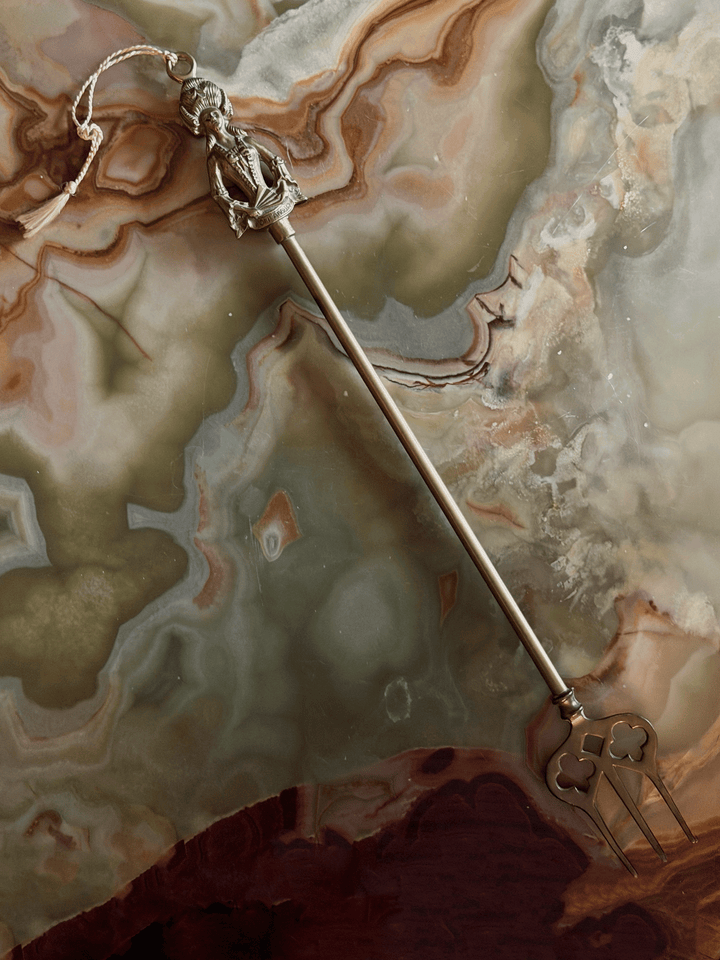 A vintage fireplace toasting fork, resting on a swirling cream and green onyx surface. The finial is sculpted into the shape of Madame de Pompadour in 18th-century French dress with a towering, high-swept hairstyle and a laced bodice. She holds a folded hand fan across her front that has the word POMPADOUR stamped clearly into it. A thin, braided cream string tassel is tied to a suspension ring at the very top of her head.
