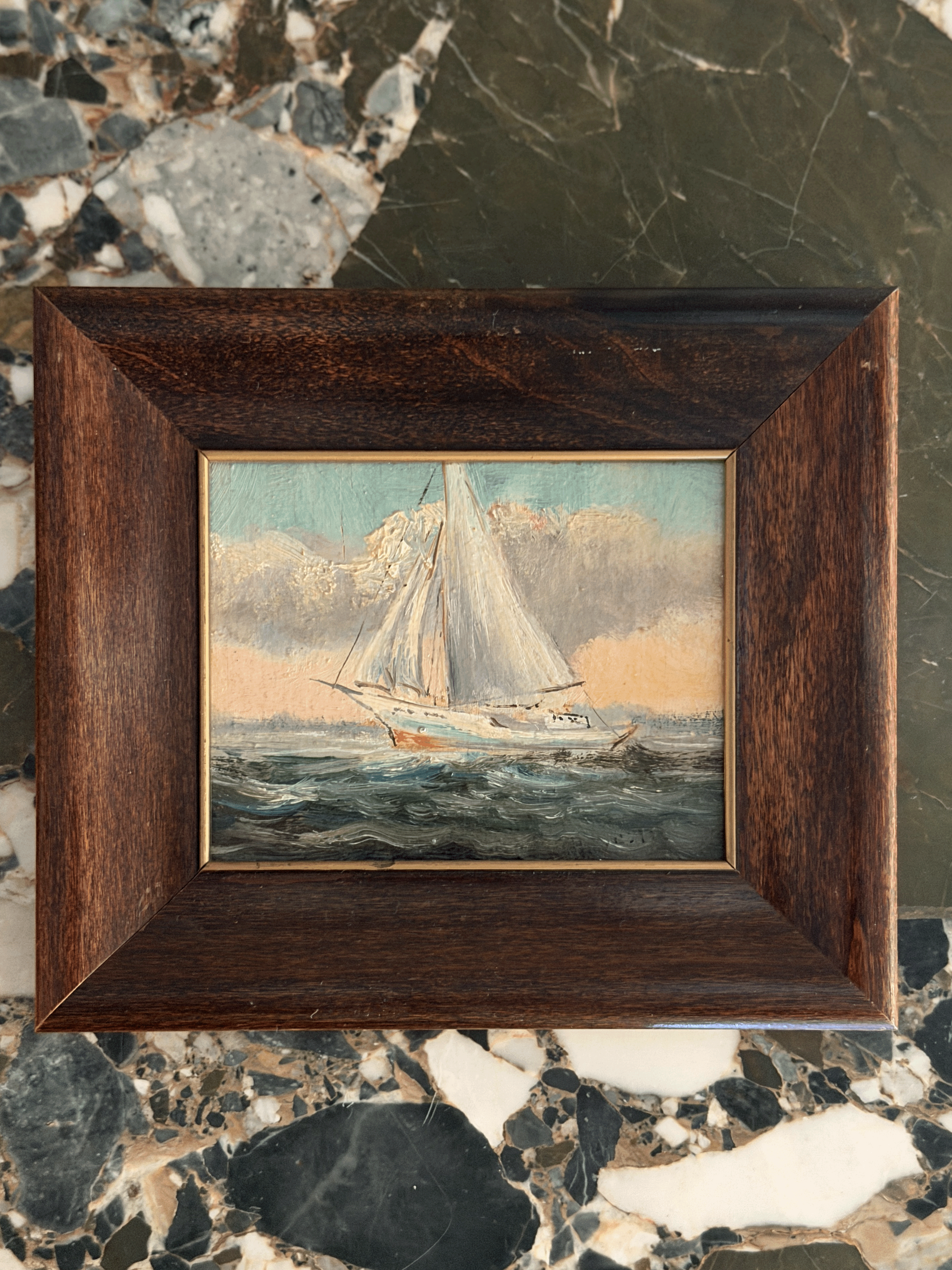 A framed miniature oil painting of a sailboat on the sea, set in a dark brown wooden frame and displayed on a polished marble surface.