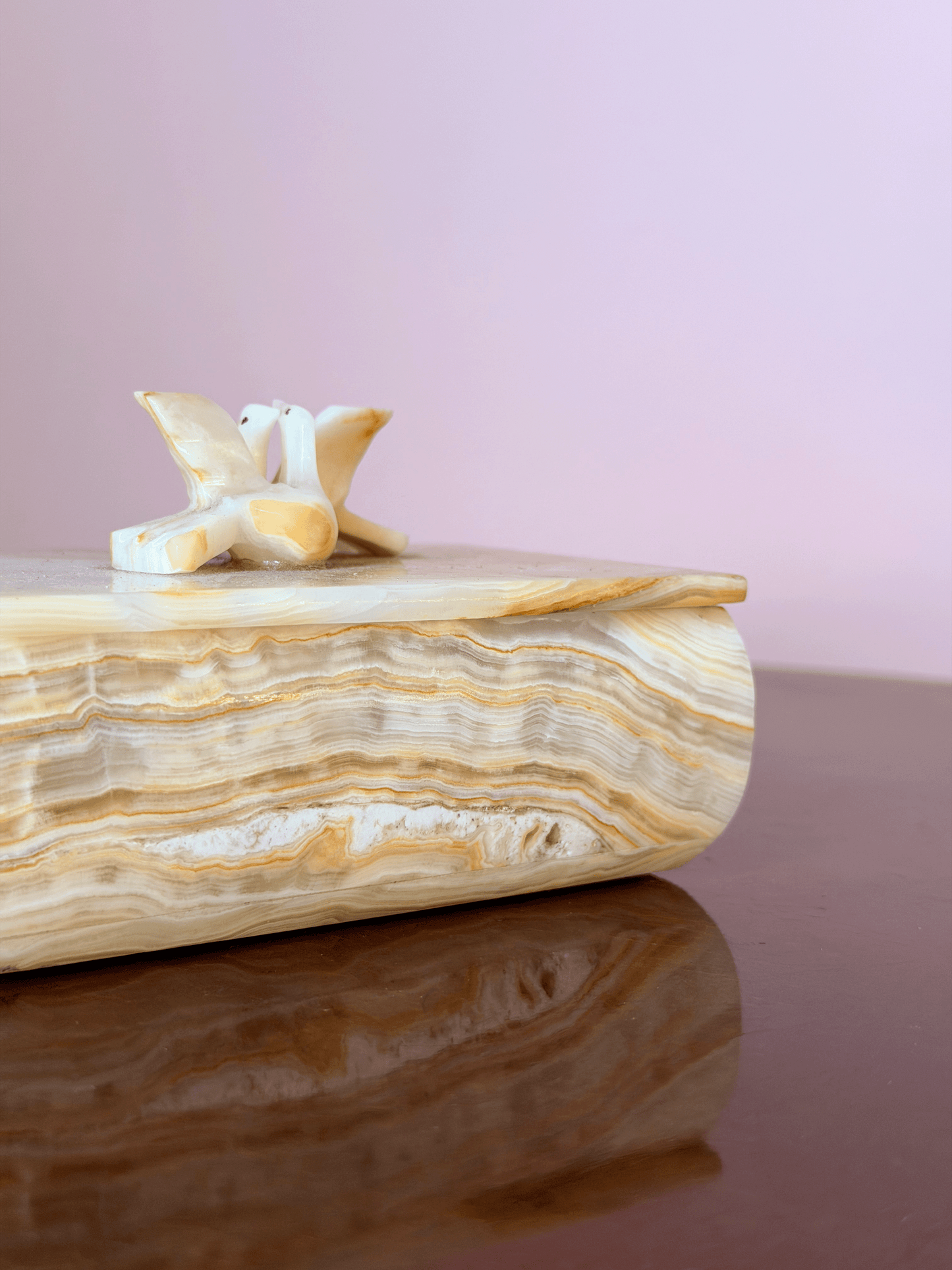 A decorative box made of polished beige and cream banded stone, featuring a small carved bird figurine on top, set against a soft pink background and dark reflective surface.