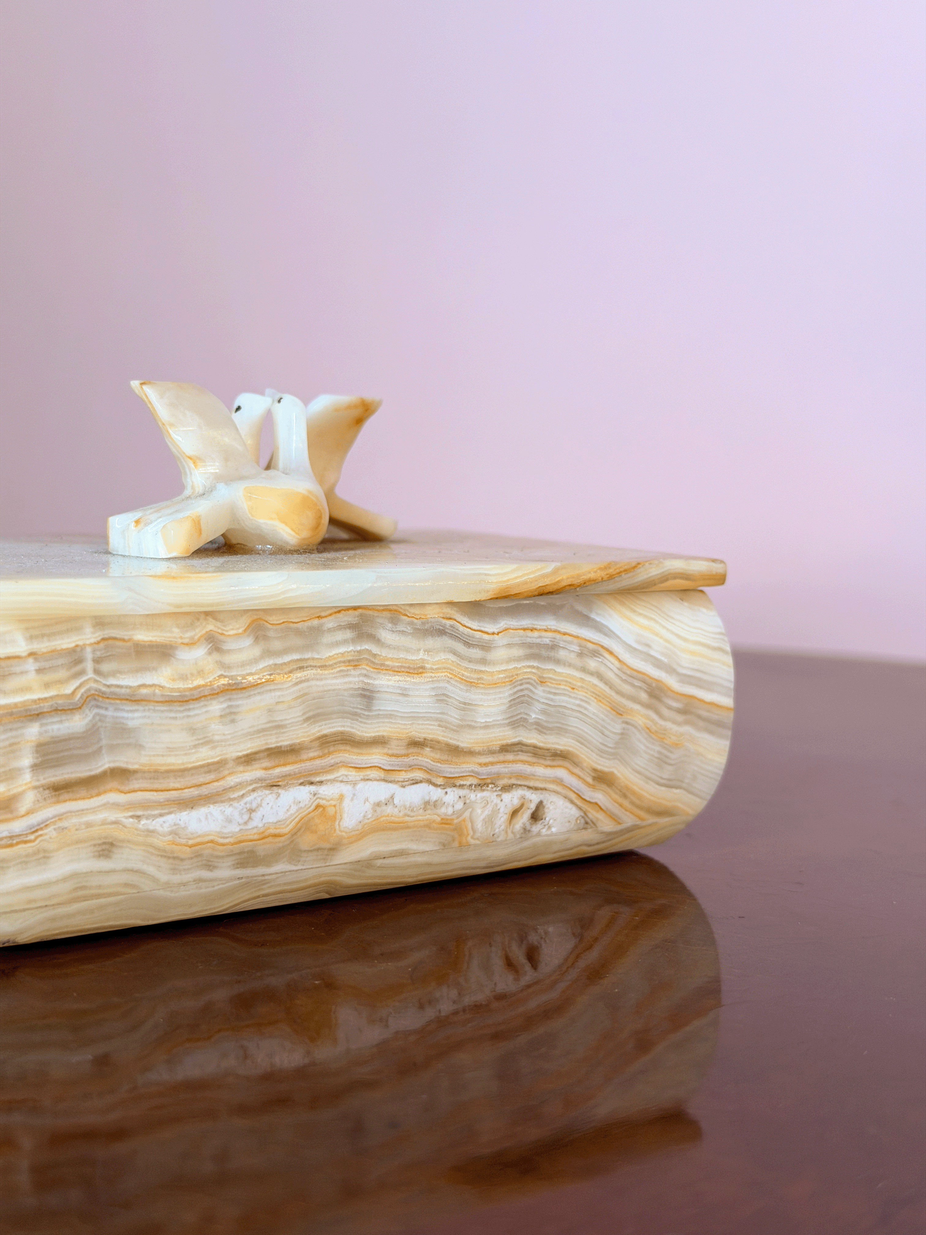 A decorative box made of polished beige and cream banded stone, featuring a small carved bird figurine on top, set against a soft pink background and dark reflective surface.