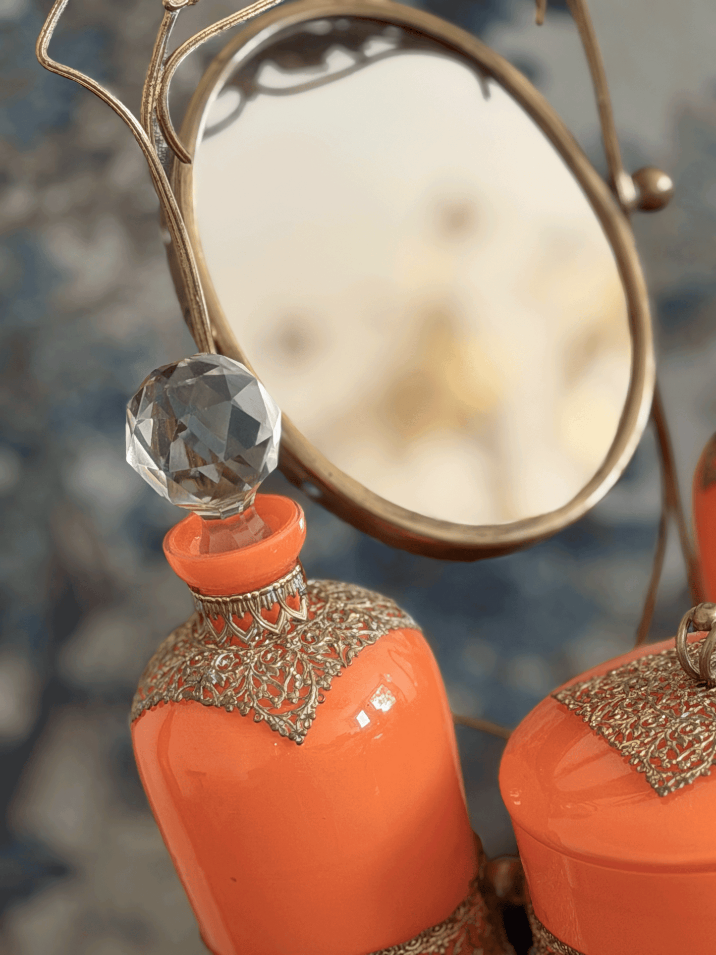 Close-up of decorative orange glass bottles with ornate silver lace-like trim and faceted clear crystal stoppers, alongside a round brass mirror.