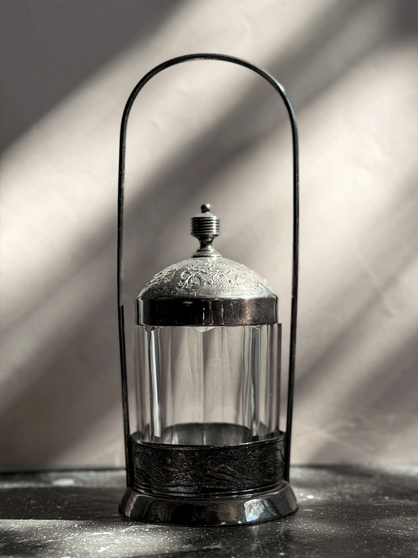 Antique Victorian silver-plated pickle castor featuring a clear vertical-paneled glass jar nested inside a tall, arched lantern-style metal frame with a shadow backdrop.