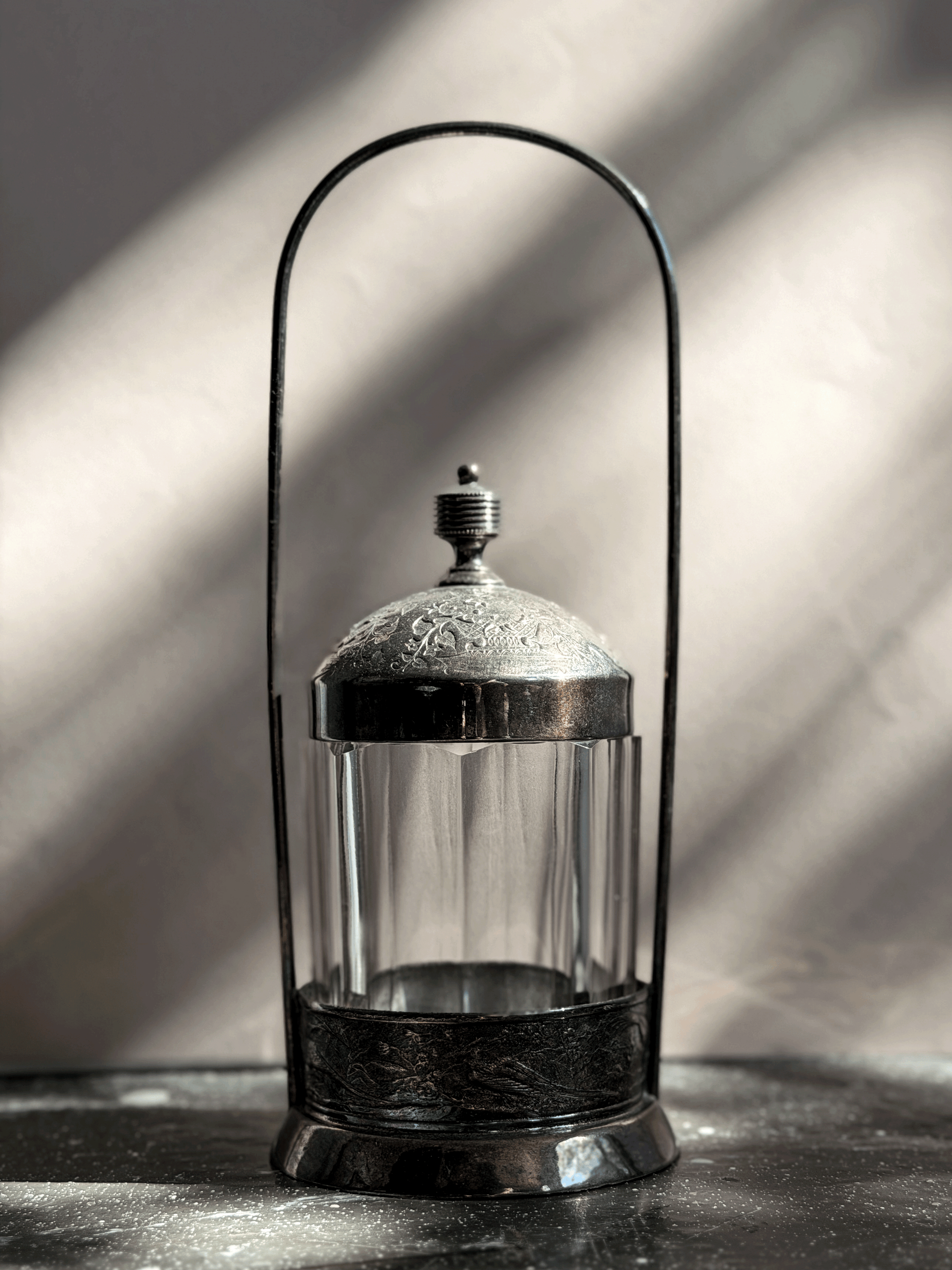 Antique Victorian silver-plated pickle castor featuring a clear vertical-paneled glass jar nested inside a tall, arched lantern-style metal frame with a shadow backdrop.