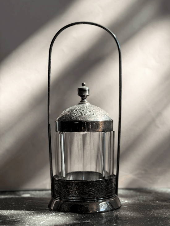 Antique Victorian silver-plated pickle castor featuring a clear vertical-paneled glass jar nested inside a tall, arched lantern-style metal frame with a shadow backdrop.