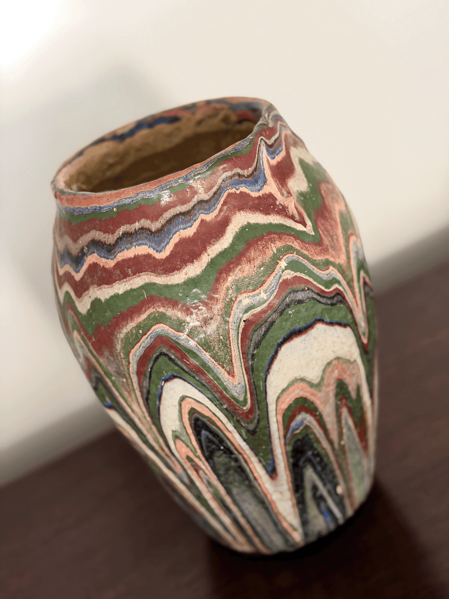 A handmade ozark pottery vase with a marbled, wavy drip glaze  pattern of green, red, blue, beige, and black, sitting on a dark wooden surface.