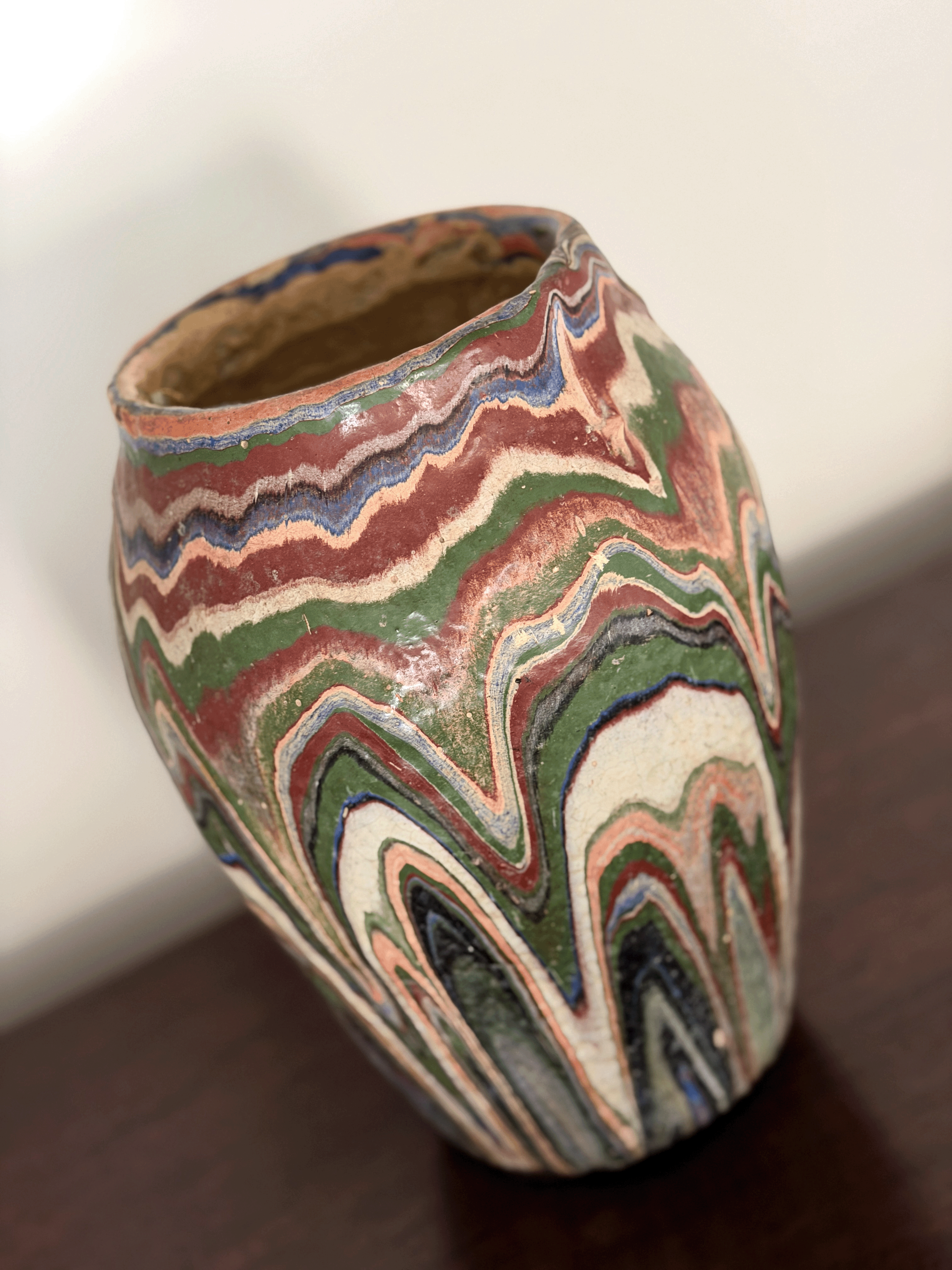A handmade ozark pottery vase with a marbled, wavy drip glaze  pattern of green, red, blue, beige, and black, sitting on a dark wooden surface.