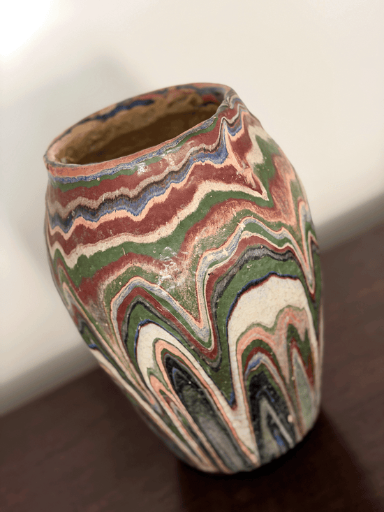 A handmade ozark pottery vase with a marbled, wavy drip glaze  pattern of green, red, blue, beige, and black, sitting on a dark wooden surface.