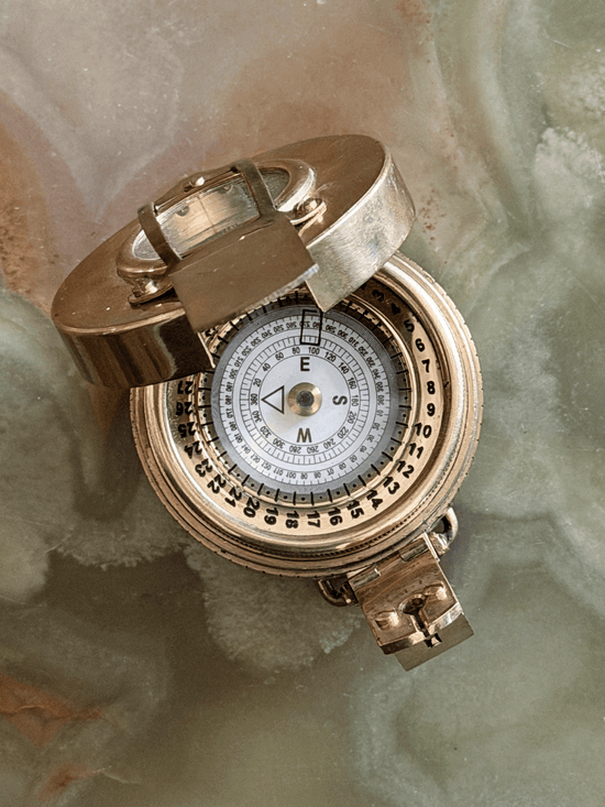 Vintage reproduction circular brass military prismatic sighting compass with a darkened antiqued patina, showing an open protective lid, a folding magnifying prism arm, and a white degree-graduated floating dial face resting on an onyx stone surface.