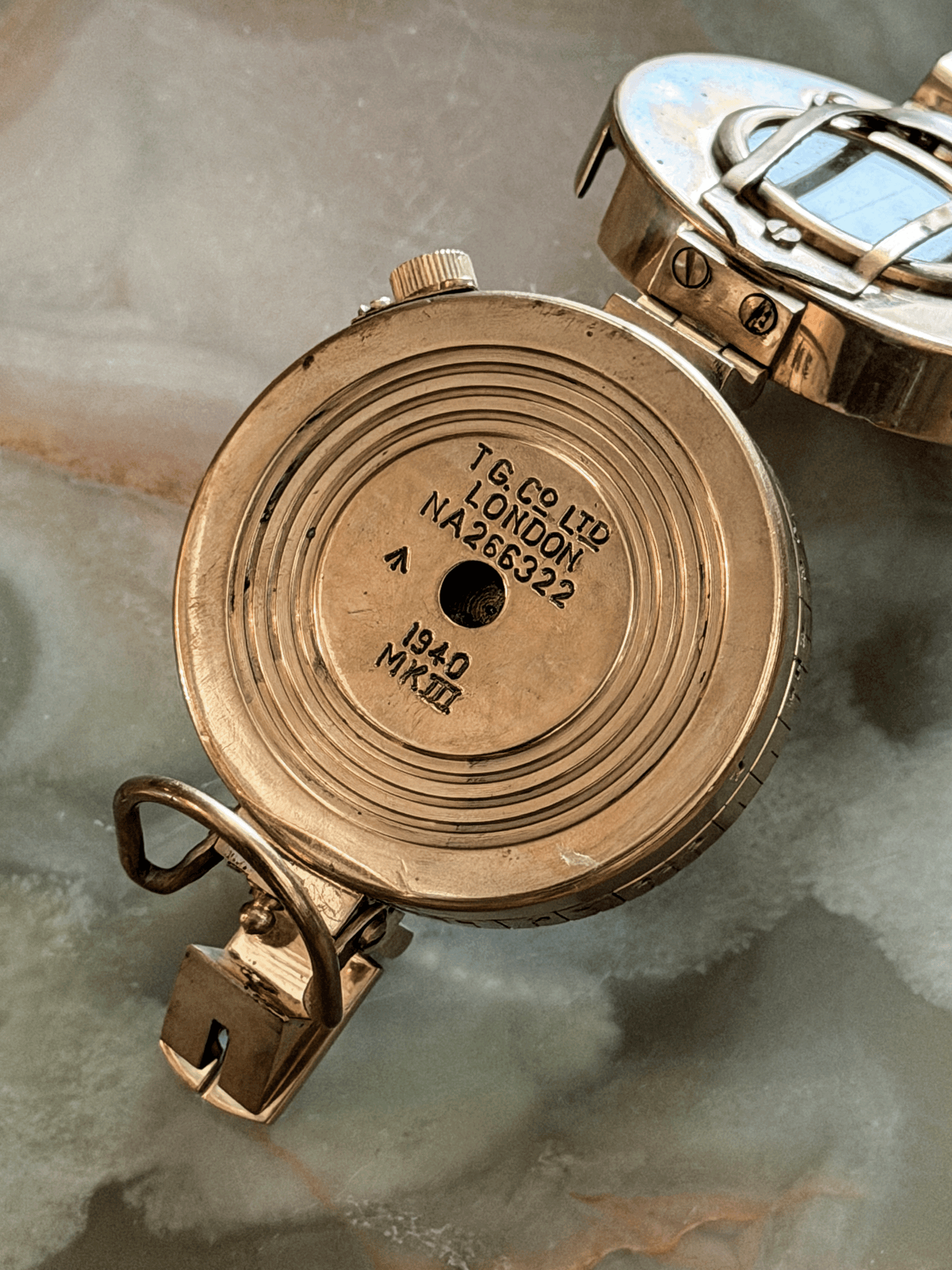 Underside base plate of a vintage reproduction brass military compass, showing crisp concentric circular engravings stamped with TG CO LTD LONDON, NA266322, 1940 MK III, and the historic British War Department Broad Arrow mark.