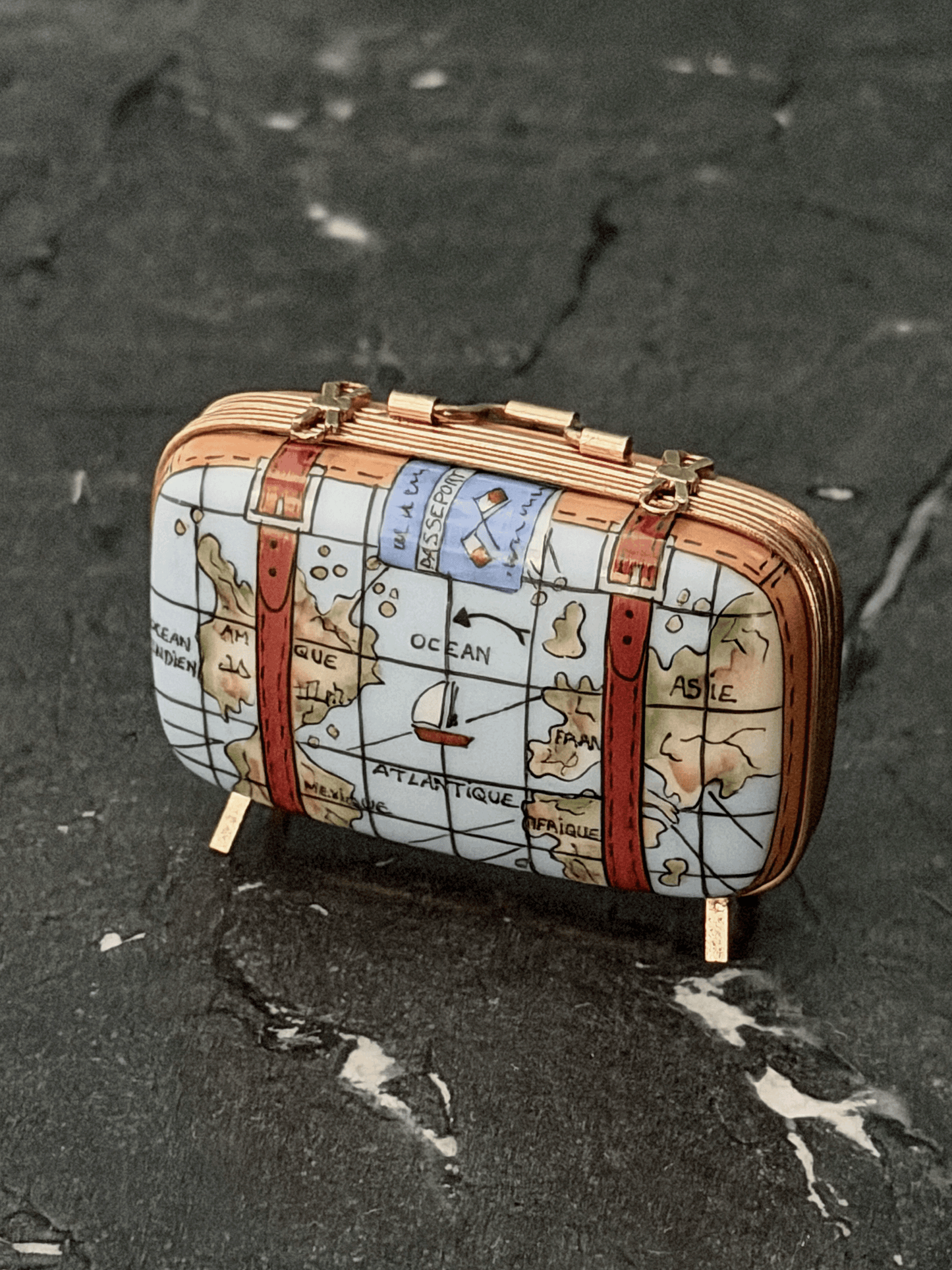 Hand-painted Rochard Limoges porcelain travel trinket box shaped like a vintage rectangular suitcase, decorated with colorful world map details, leather corner reinforcement plates, luggage straps with gold buckles, and standing on four brass peg feet.