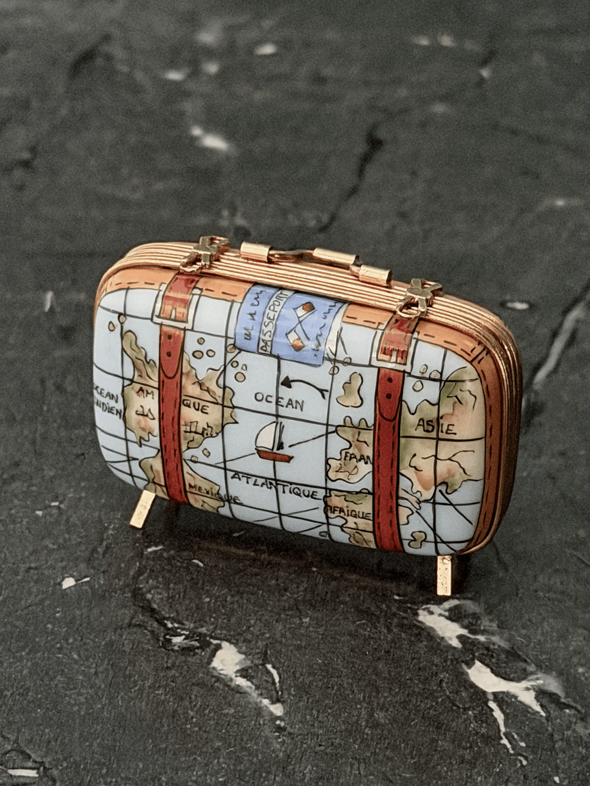 Hand-painted Rochard Limoges porcelain travel trinket box shaped like a vintage rectangular suitcase, decorated with colorful world map details, leather corner reinforcement plates, luggage straps with gold buckles, and standing on four brass peg feet.