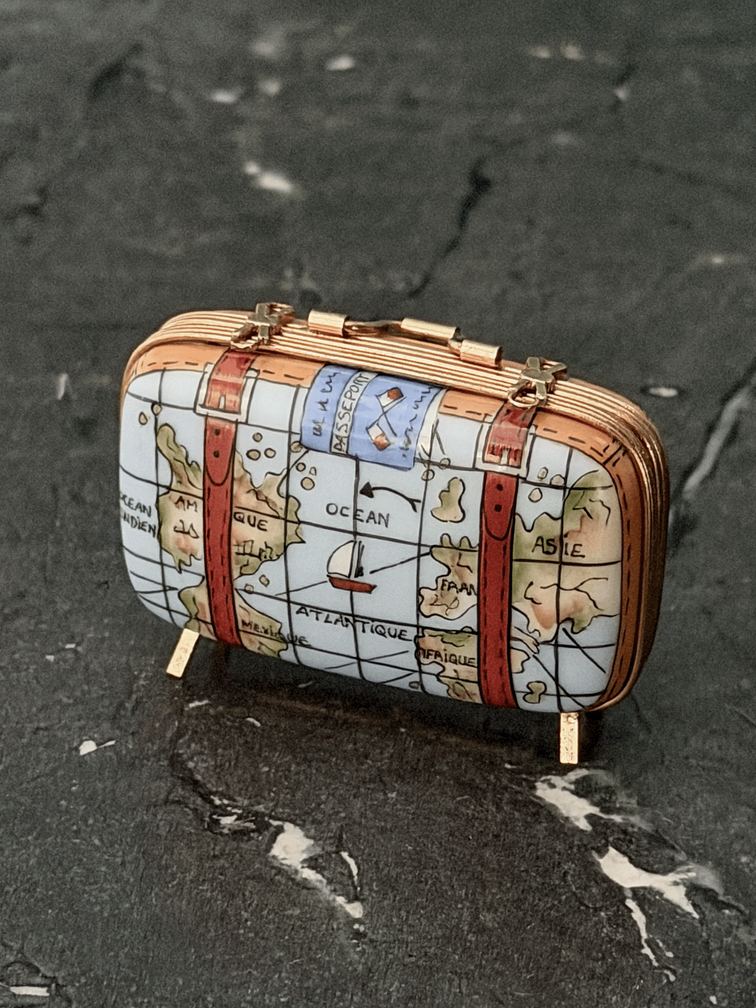 Hand-painted Rochard Limoges porcelain travel trinket box shaped like a vintage rectangular suitcase, decorated with colorful world map details, leather corner reinforcement plates, luggage straps with gold buckles, and standing on four brass peg feet.