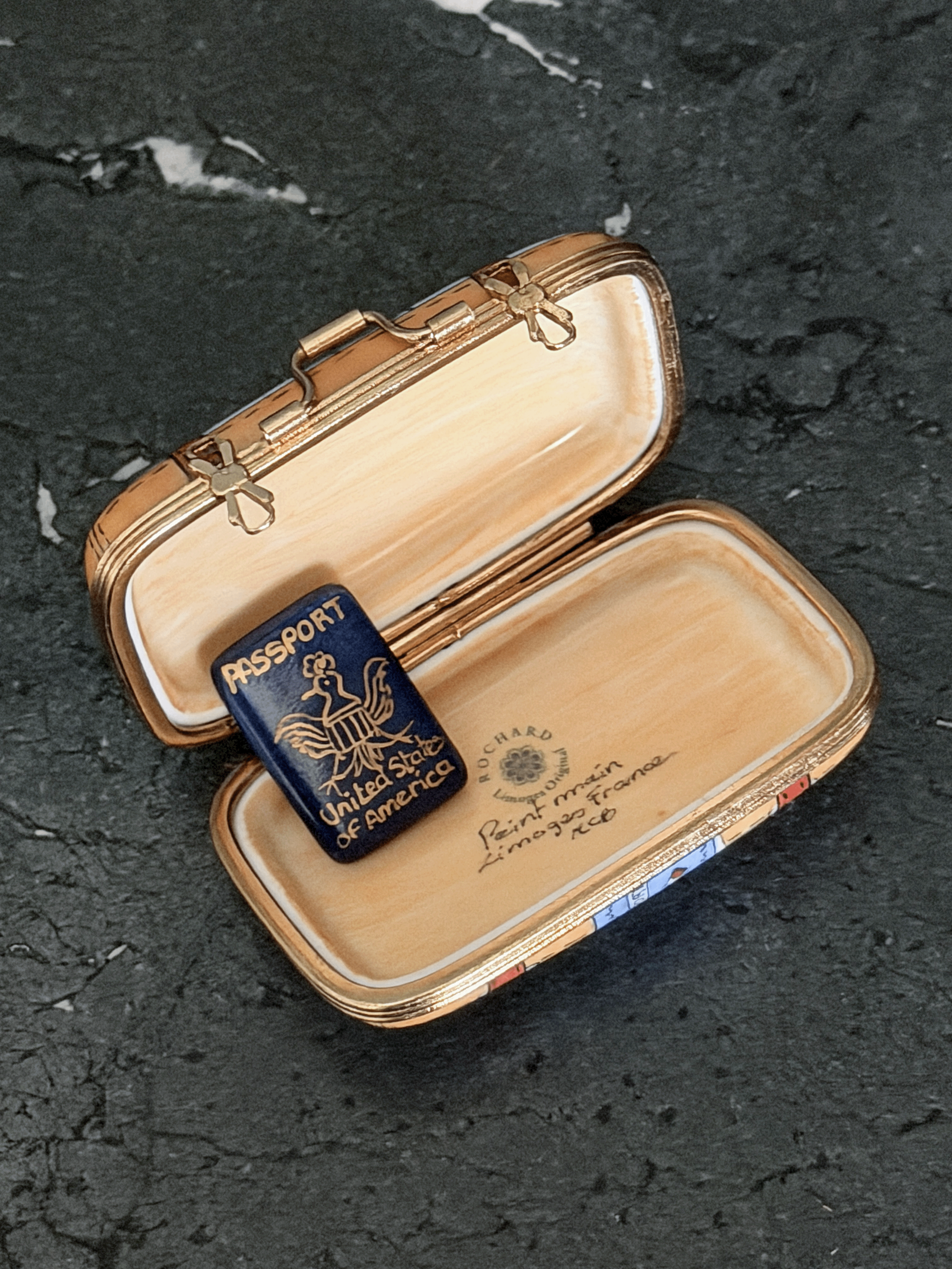 Opened interior of a Rochard map suitcase Limoges box showing a tan leather textile texture lining, the factory original workshop backstamp, and a separate loose removable miniature navy blue porcelain United States Passport accessory with a gold bald eagle seal.