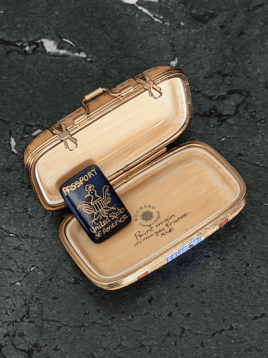 Opened interior of a Rochard map suitcase Limoges box showing a tan leather textile texture lining, the factory original workshop backstamp, and a separate loose removable miniature navy blue porcelain United States Passport accessory with a gold bald eagle seal.