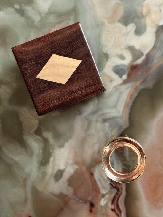 A square dark wood box with a light wood diamond inlay sits on a marbled stone surface, alongside a small round brass map magnifier 