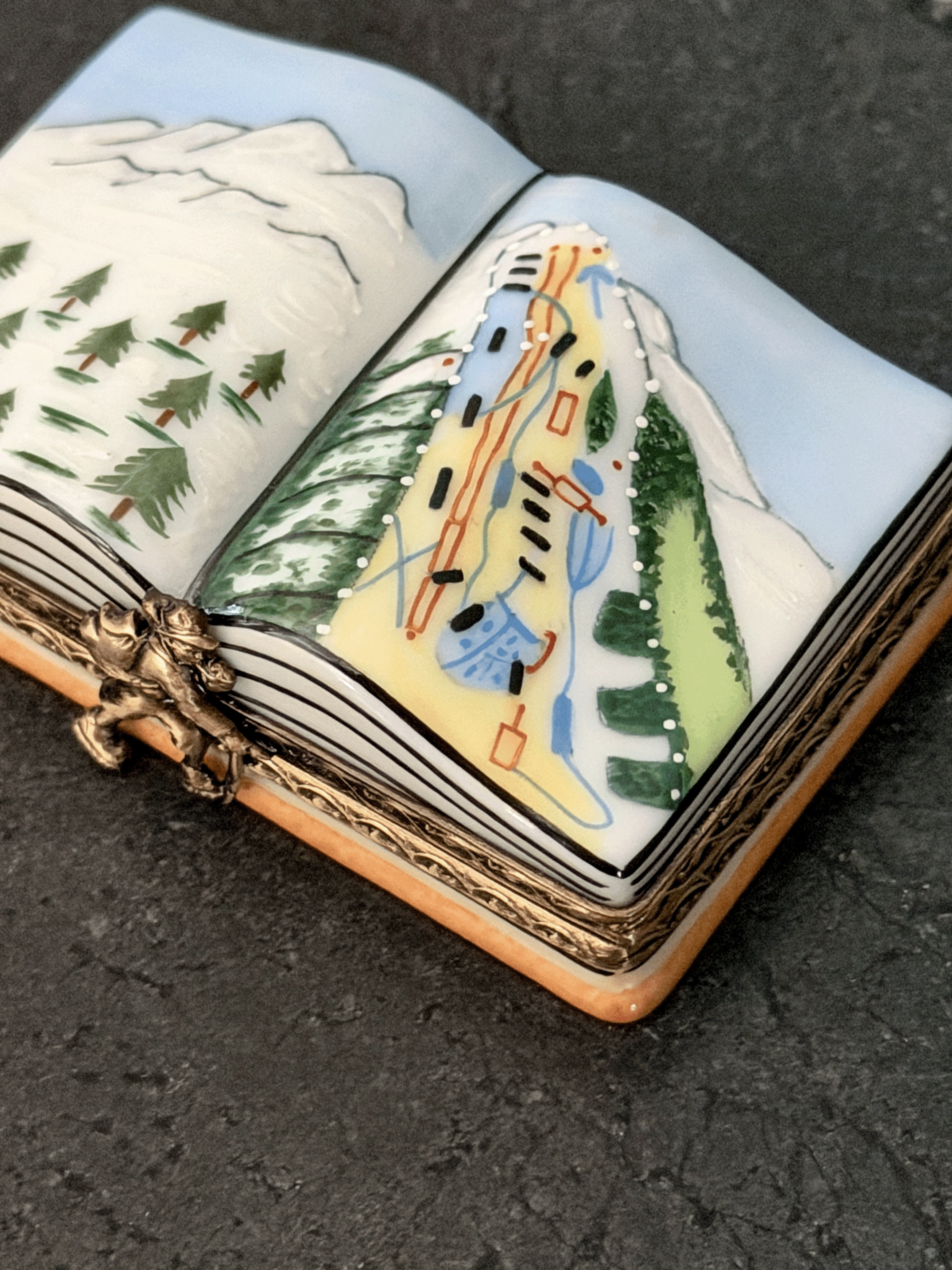 Hand-painted Rochard Limoges porcelain trinket box shaped like an open book, depicting a snow-capped alpine mountain peak on the left page and a colorful ski resort trail map with chairlift networks on the right page.