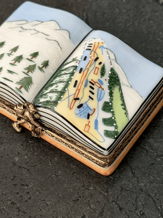 Hand-painted Rochard Limoges porcelain trinket box shaped like an open book, depicting a snow-capped alpine mountain peak on the left page and a colorful ski resort trail map with chairlift networks on the right page.