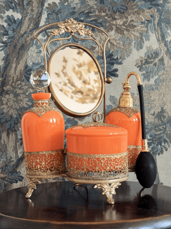 A straight-on eye-level view of a rare vintage 3-piece Art Deco Czechoslovakian orange tango glass vanity set nested in an ornate gold metal filigree caddy on a dark table. In the center is a round lidded powder jar, flanked by a cologne decanter with a large faceted crystal ball stopper on the left, and a perfume atomizer with a black woven squeeze bulb on the right. An ornate oval vanity mirror swings on a tall gold frame above the center jar. The background is a blue and tan tree-patterned tapestry.