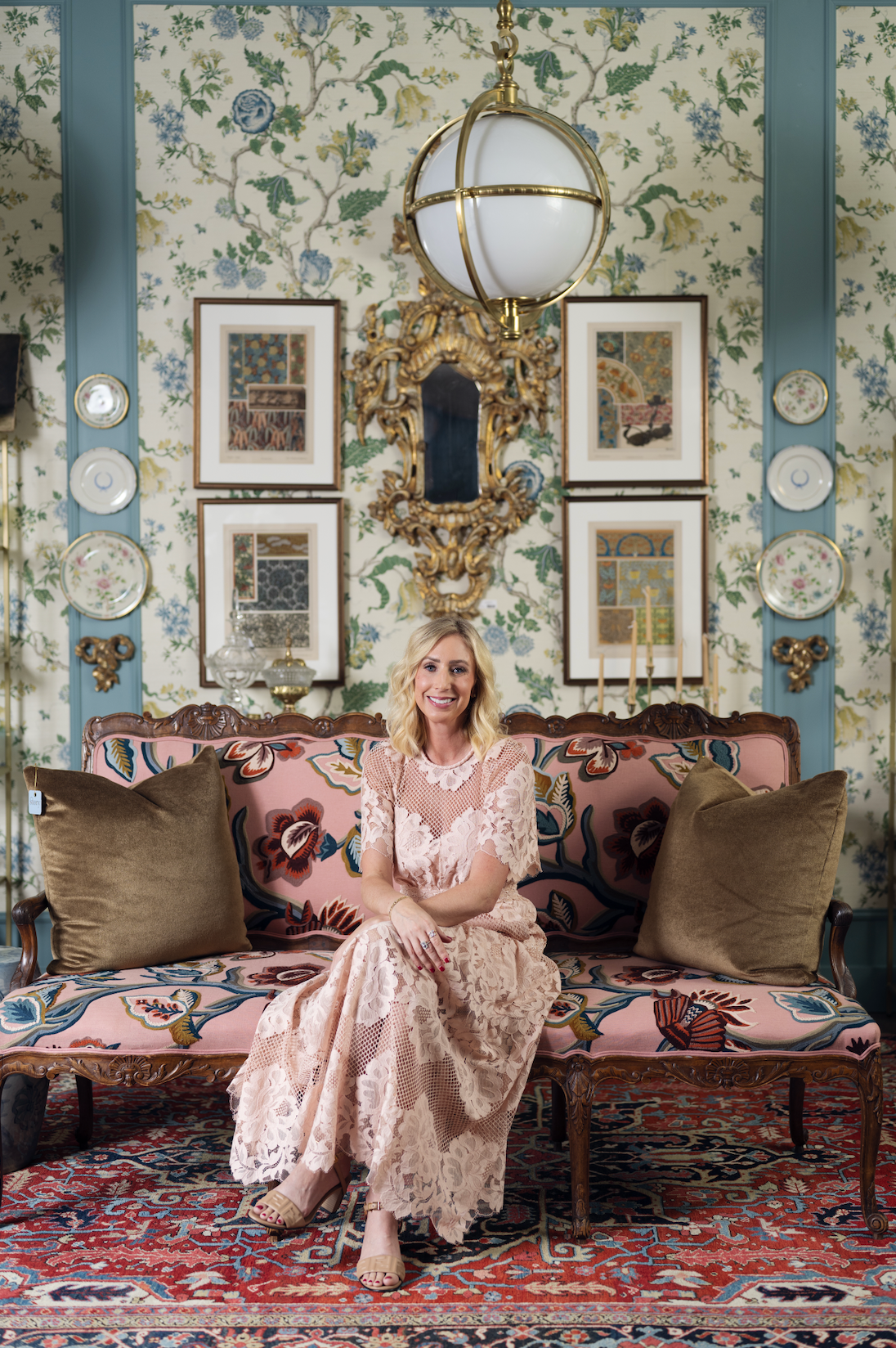 A woman sits on an ornate floral sofa in a decorated room with patterned wallpaper, framed art, a hanging globe light, and a large rug. She is wearing a pale pink lace dress.