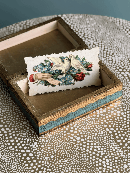A small open wooden keepsake box with distressed blue and gold painted edges, holding a vintage-style illustrated card featuring two white doves and red flowers, set on a patterned surface.