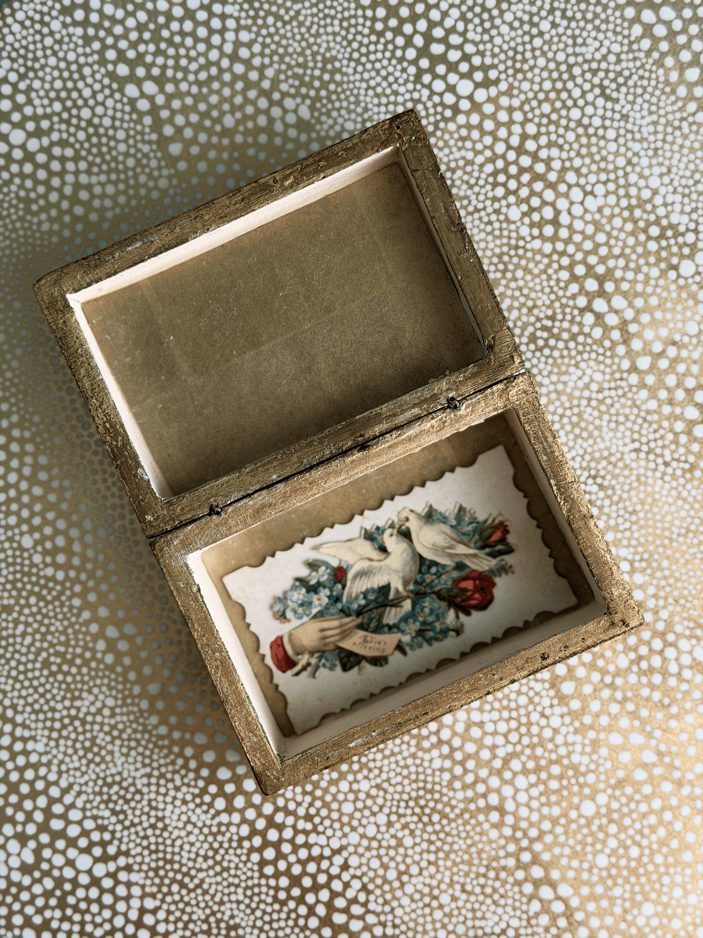 An open vintage-style wooden keepsake box with a gold-toned frame, showcasing a floral and bird illustration on the inner lining, set against a patterned beige and white background.