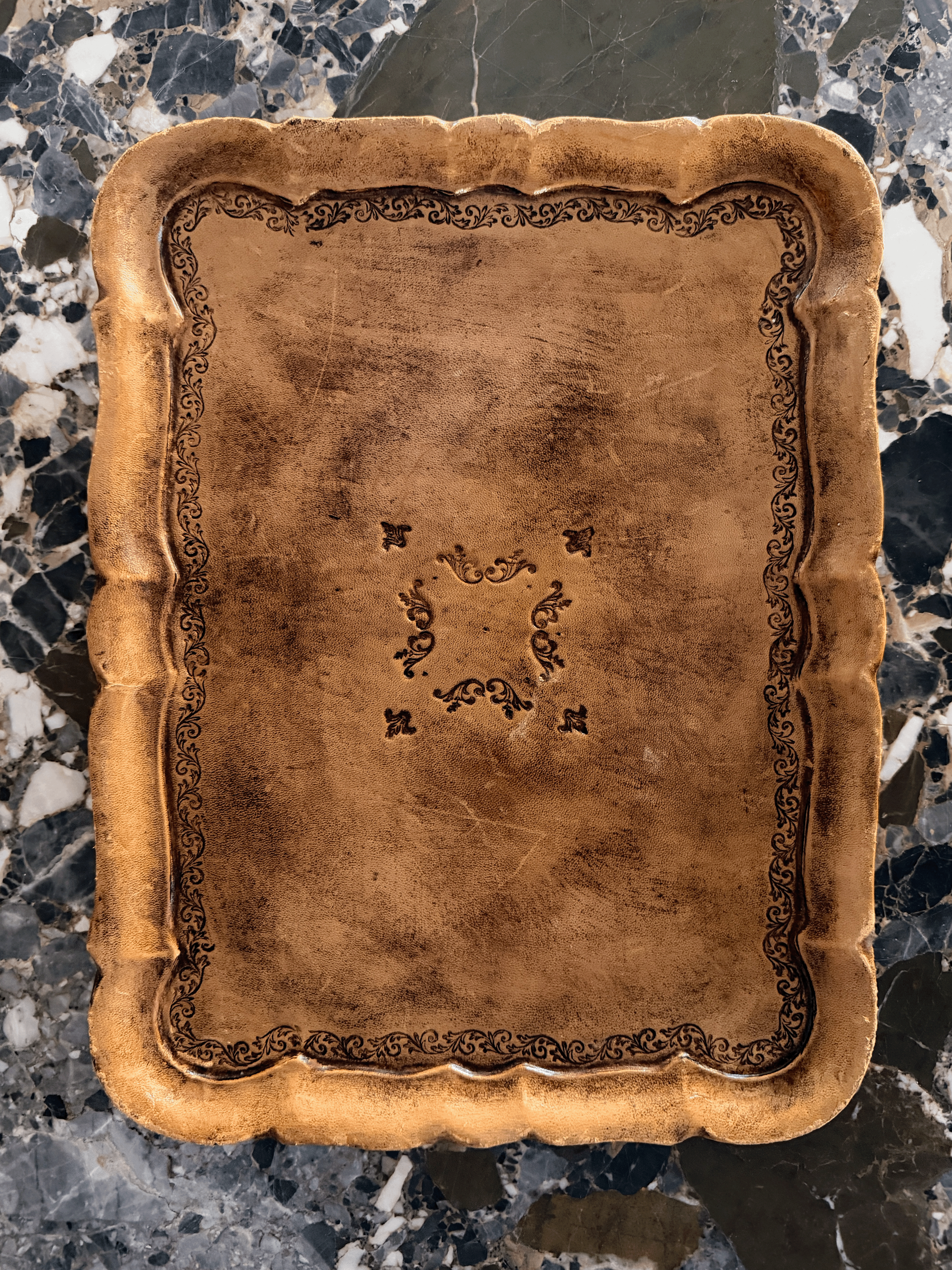 A square decorative tray with a scalloped rim and embossed floral border detailing, shown from a top-down view on a dark marble surface.