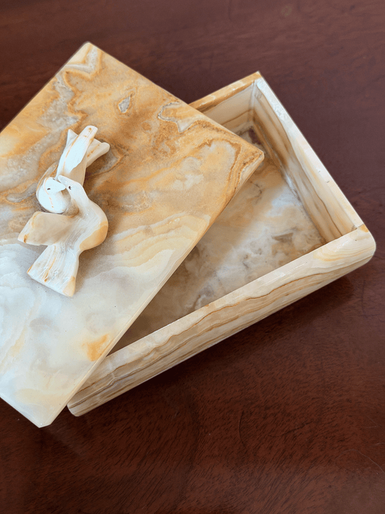 A rectangular marble keepsake box with a matching lid, featuring carved doves on top, set on a dark brown wooden surface.
