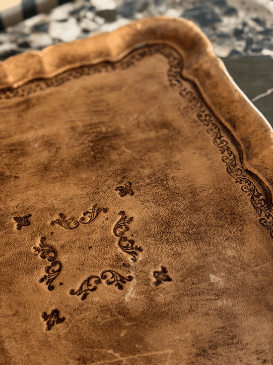 Close-Up of mid-century vintage Italian Florentine toleware serving tray wrapped in caramel-toned leather with an antiqued woodgrain patina, featuring a scalloped pie-crust rim and hand-stamped black Renaissance revival scrolling filigree.