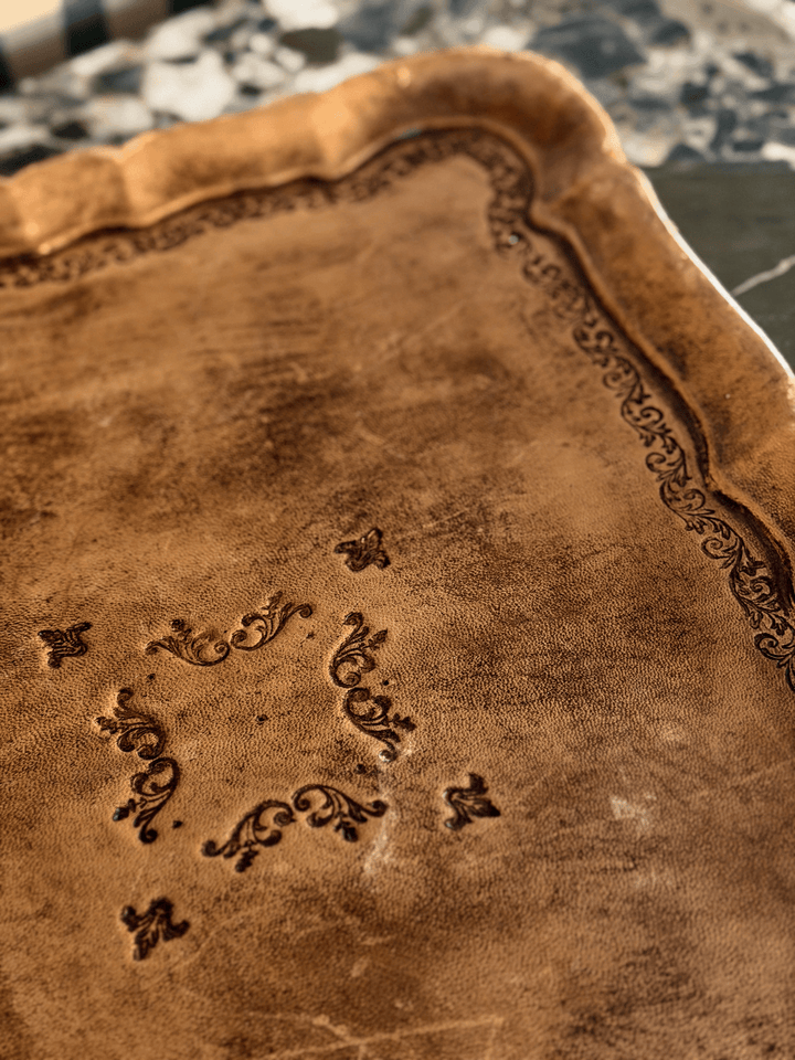 Close-Up of mid-century vintage Italian Florentine toleware serving tray wrapped in caramel-toned leather with an antiqued woodgrain patina, featuring a scalloped pie-crust rim and hand-stamped black Renaissance revival scrolling filigree.