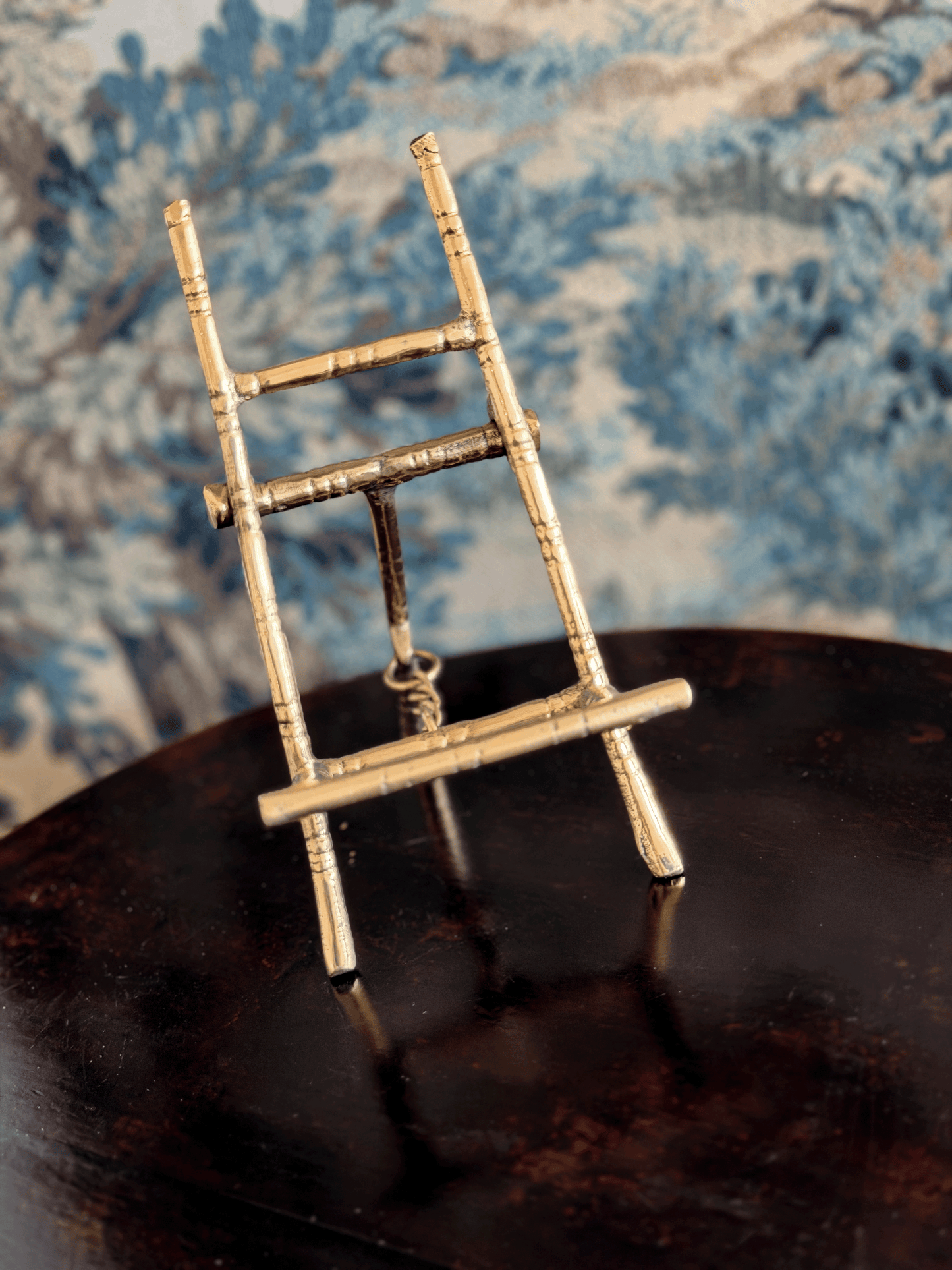 A miniature brass easel displayed on a dark reflective surface with a blue patterned fabric background.