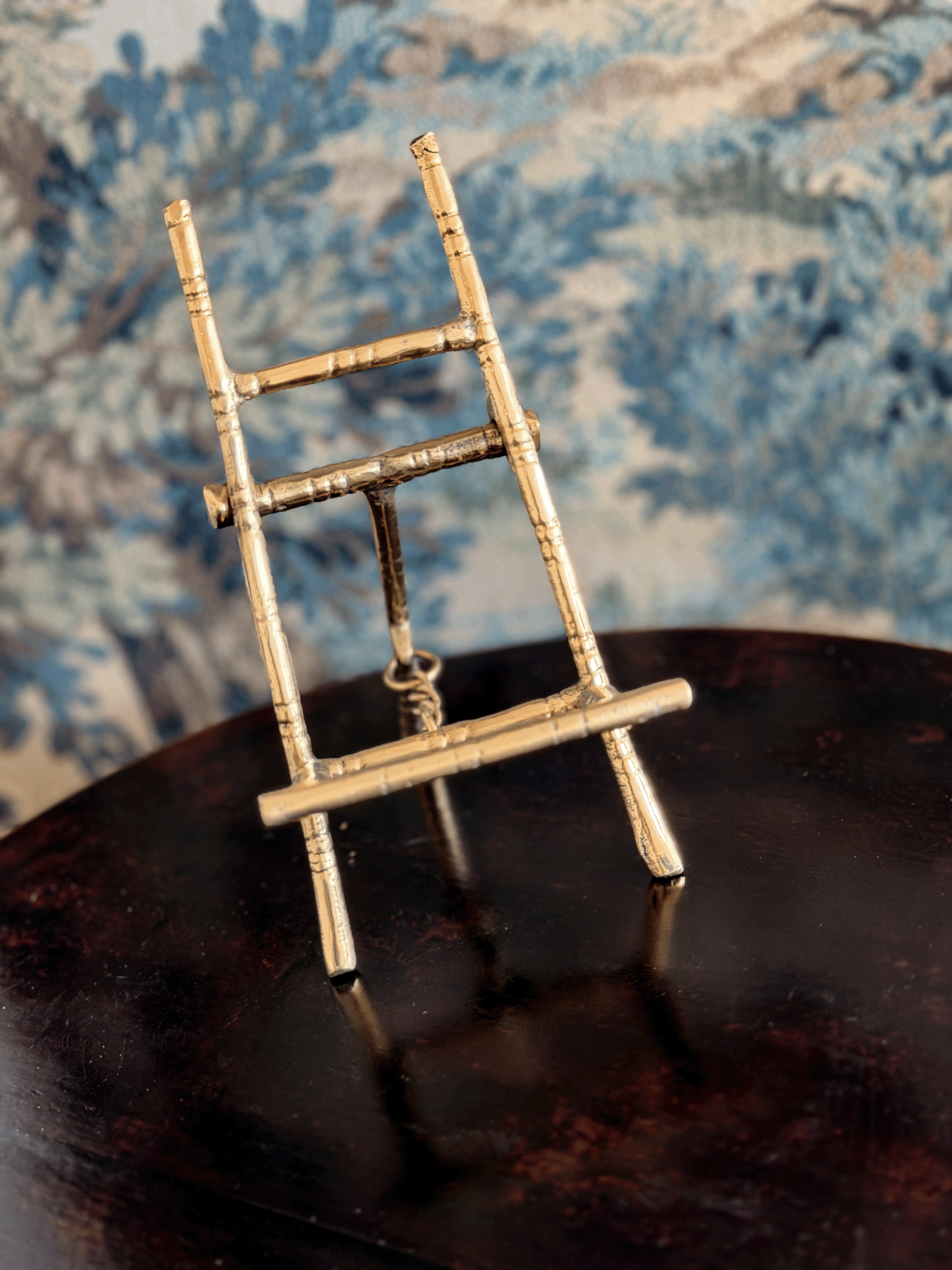A miniature brass easel displayed on a dark reflective surface with a blue patterned fabric background.