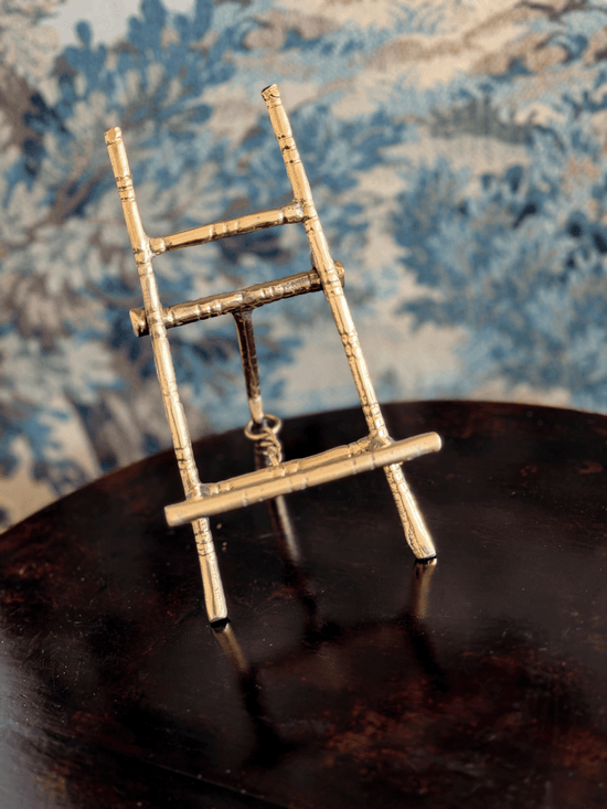 A miniature brass easel displayed on a dark reflective surface with a blue patterned fabric background.