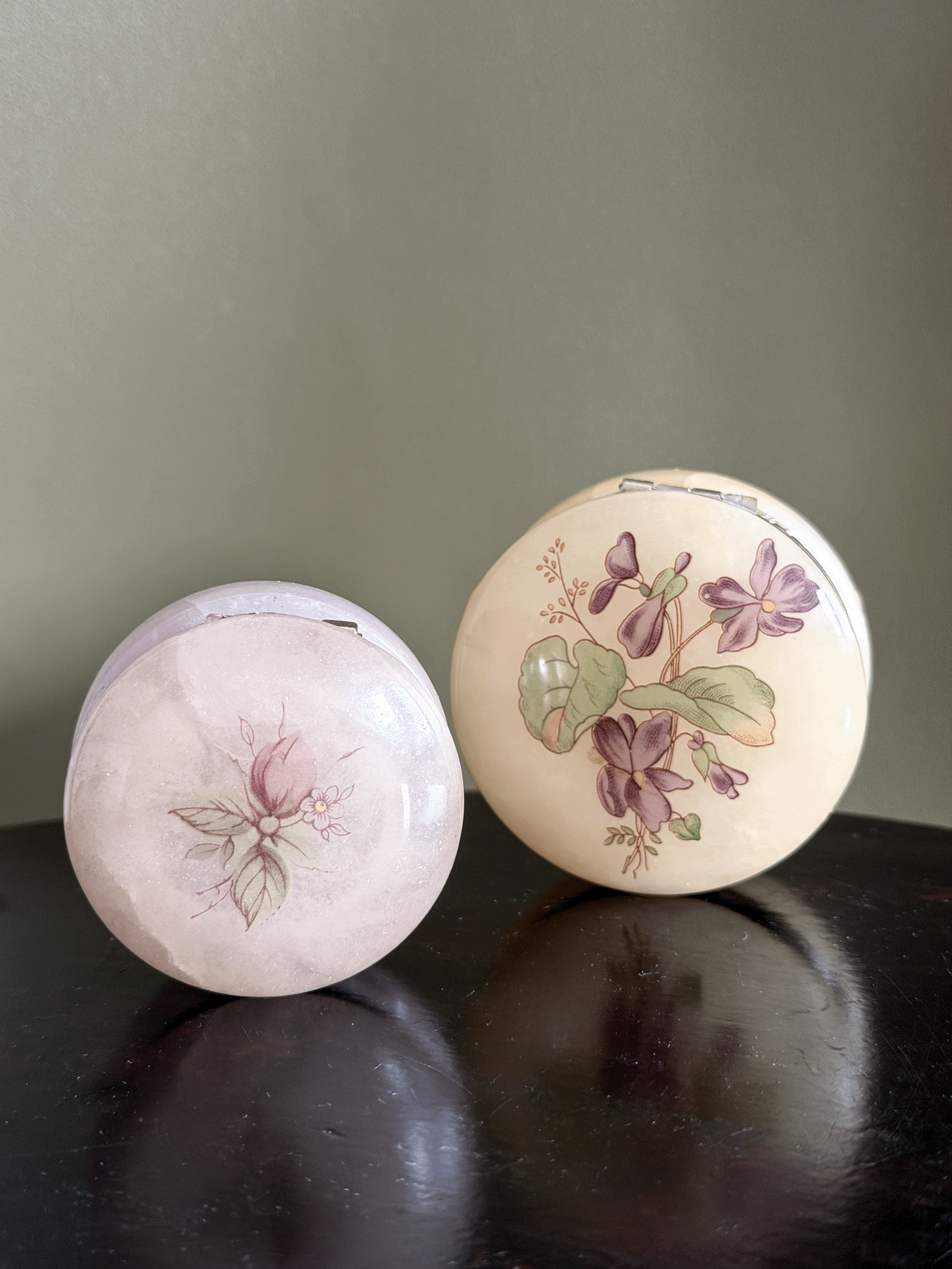 Two round vintage compact cases with floral illustrations, one pale pink and one cream with purple and pink flowers, displayed on a dark wooden surface.