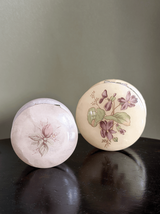 Two round vintage compact cases with floral illustrations, one pale pink and one cream with purple and pink flowers, displayed on a dark wooden surface.