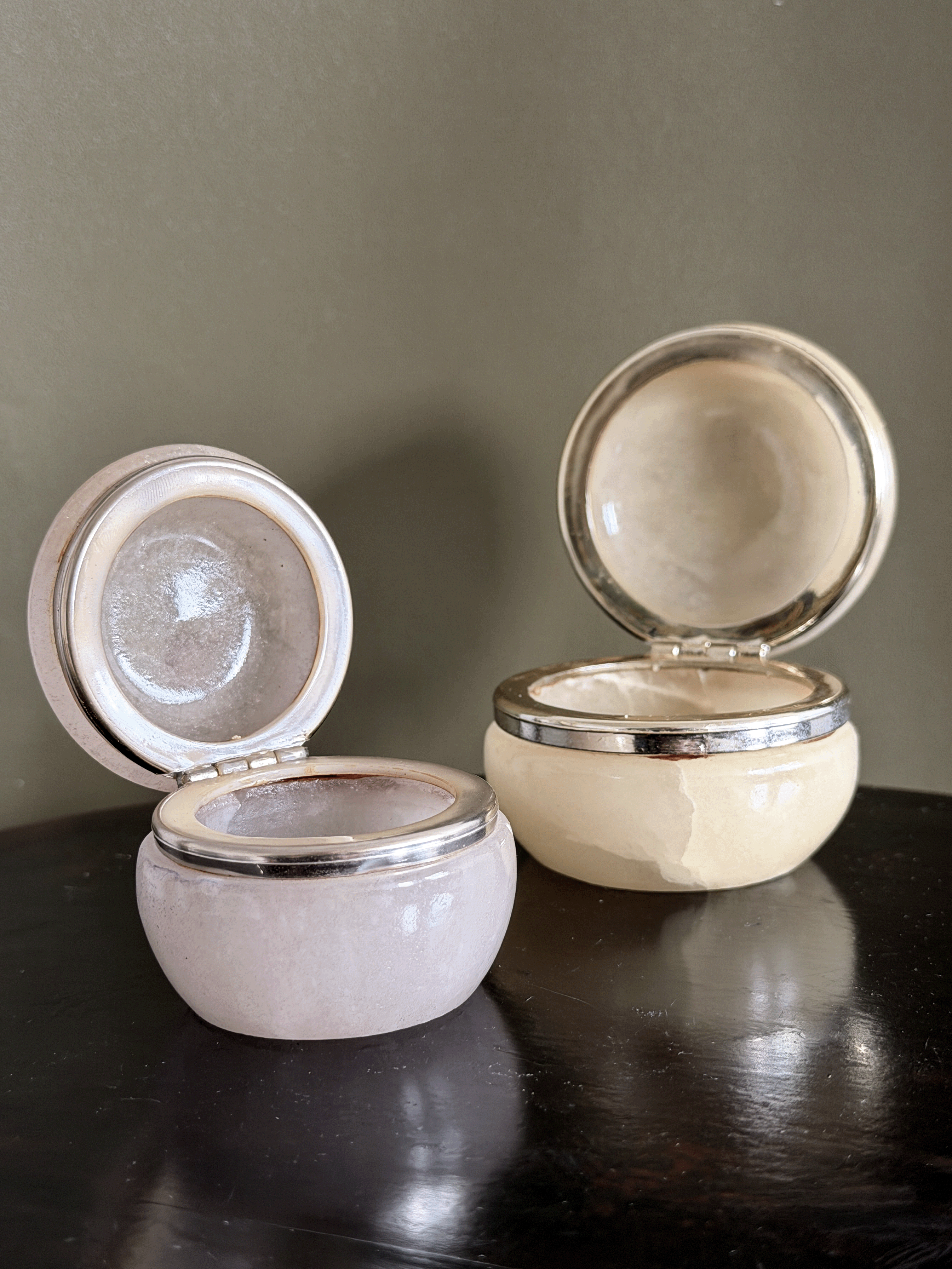 Two small round genuine Italian alabaster carved vanity jars, shown open on a dark reflective surface against a muted olive-gray background.