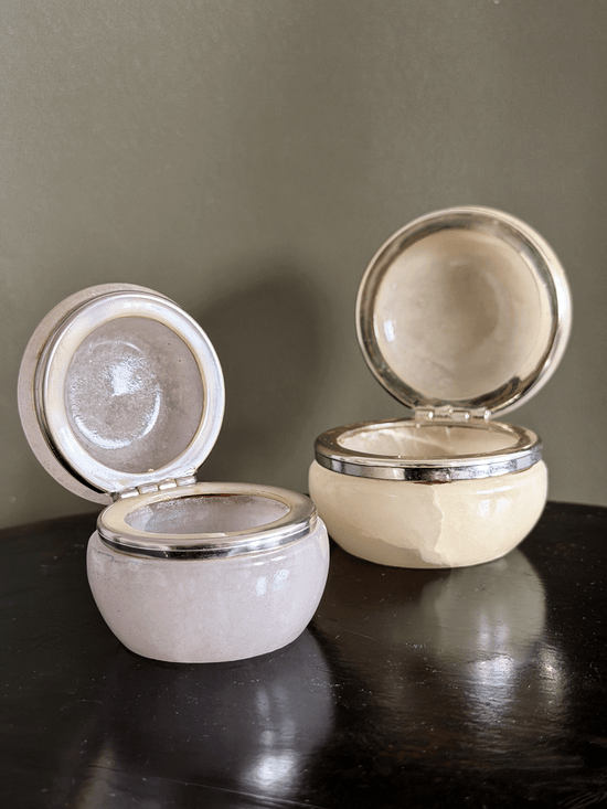 Two small round genuine Italian alabaster carved vanity jars, shown open on a dark reflective surface against a muted olive-gray background.